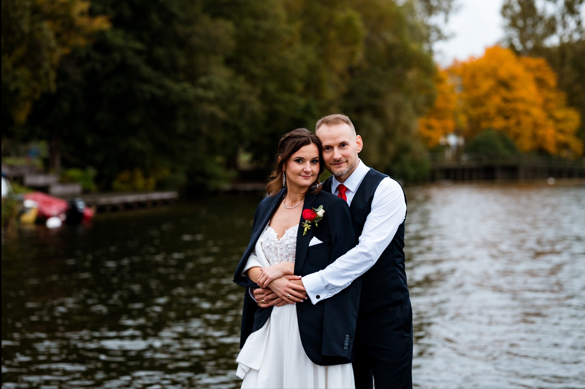 Para młoda w eleganckich strojach pozuje na tle jeziora i jesiennych drzew. Pan obejmuje Pannę Młodą, oboje uśmiechnięci, widać obrączki. Romantyczna sceneria.