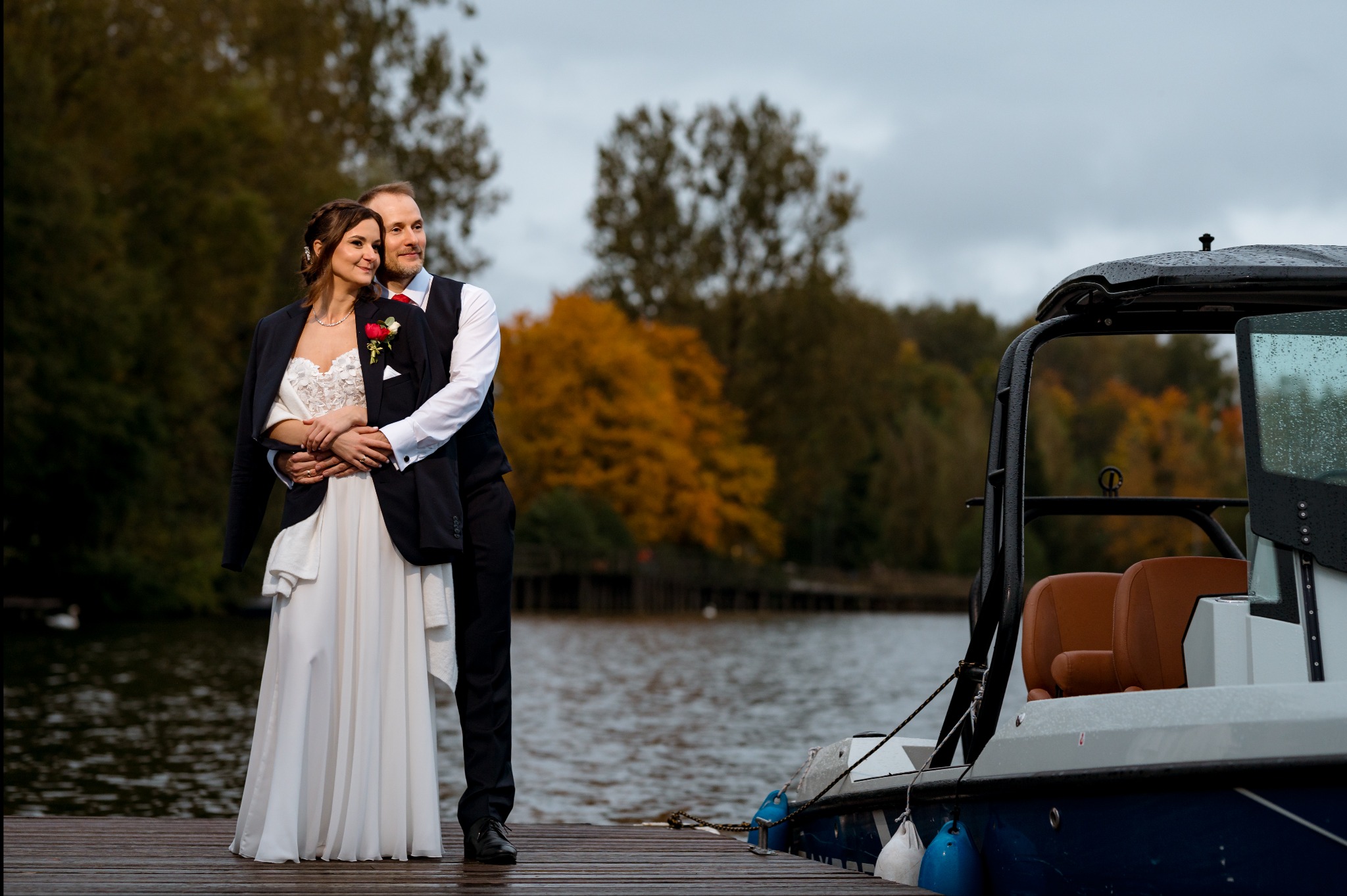 Para młoda na drewnianym pomoście, w tle woda i jesienne drzewa. Uwagę przykuwa nowoczesna łódź motorowa po prawej stronie kadru. Romantyczna sesja ślubna.