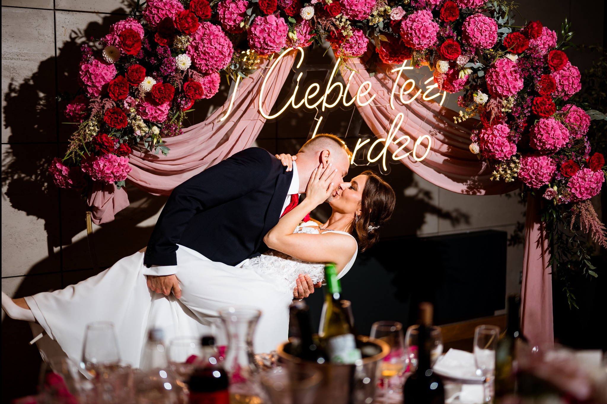 Panna młoda w białej sukni, unoszona przez pana młodego w ciemnym garniturze, całuje się na tle kwiatowej dekoracji z neonowym napisem.