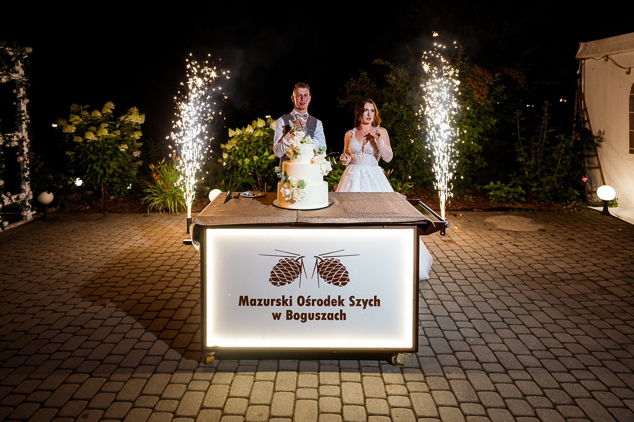 Uśmiechnięta para młoda kroi tort weselny na tle fontanny iskier, na zewnątrz, nocą. Stół z podświetlanym logo 'Mazurski Ośrodek Szych w Boguszach'.