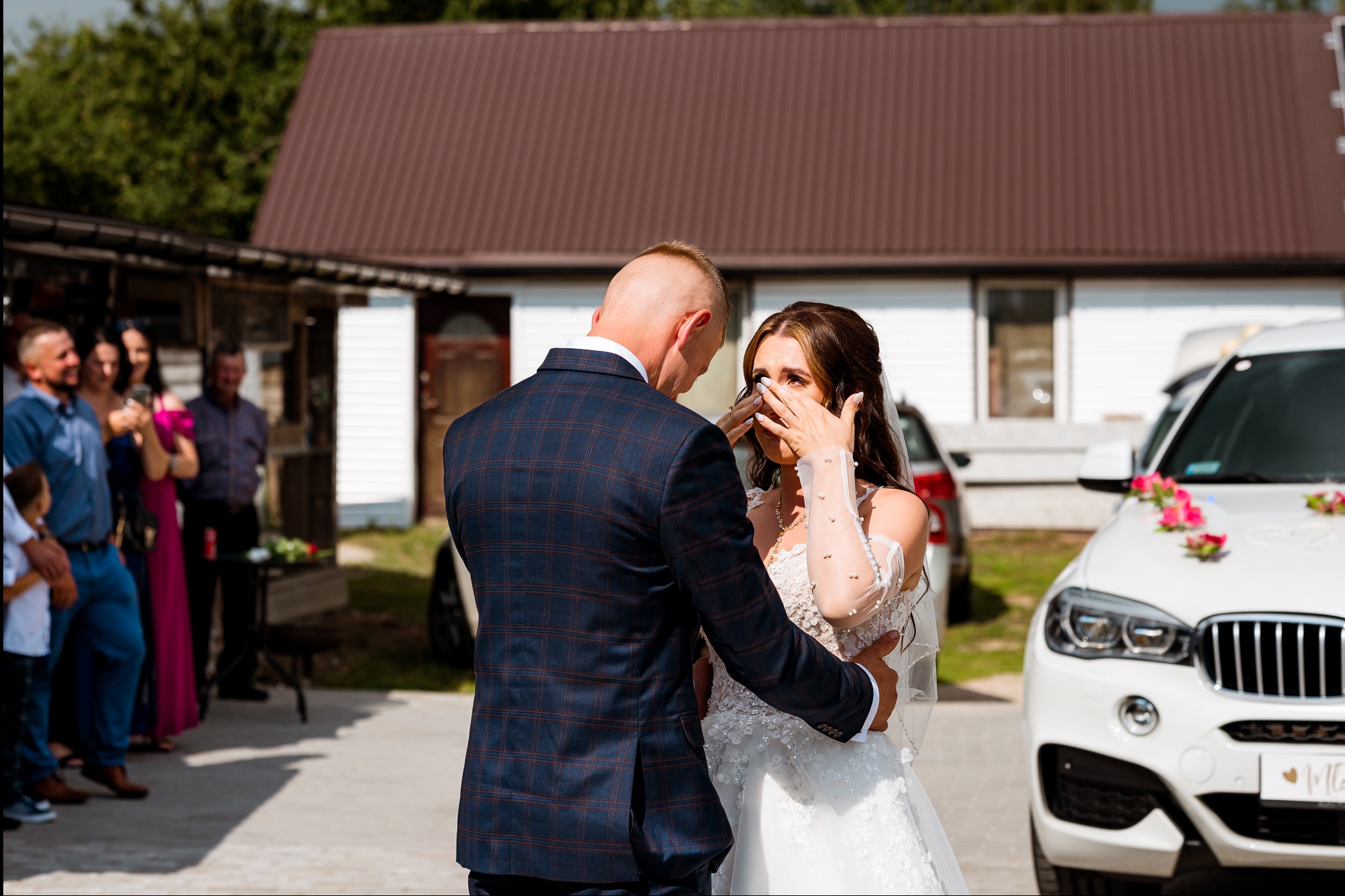 Wzruszona panna młoda w objęciach pana młodego, na tle udekorowanego samochodu i gości weselnych. Ujęcie z ceremonii ślubnej w plenerze, w słoneczny dzień.