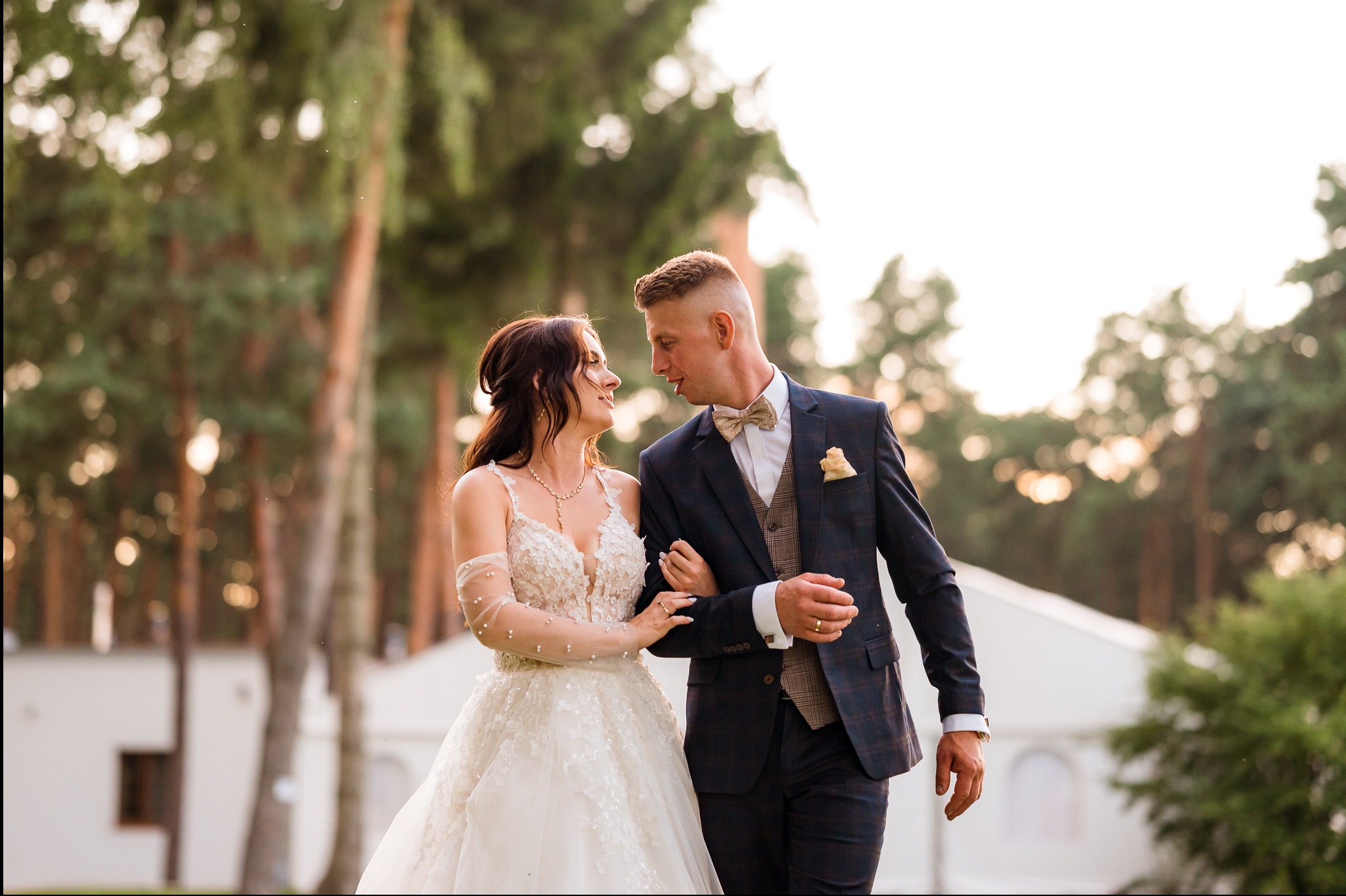 Uśmiechnięta para młoda w plenerze: panna młoda w koronkowej sukni i pan młody w eleganckim garniturze, spacerują trzymając się za ręce na tle zieleni.