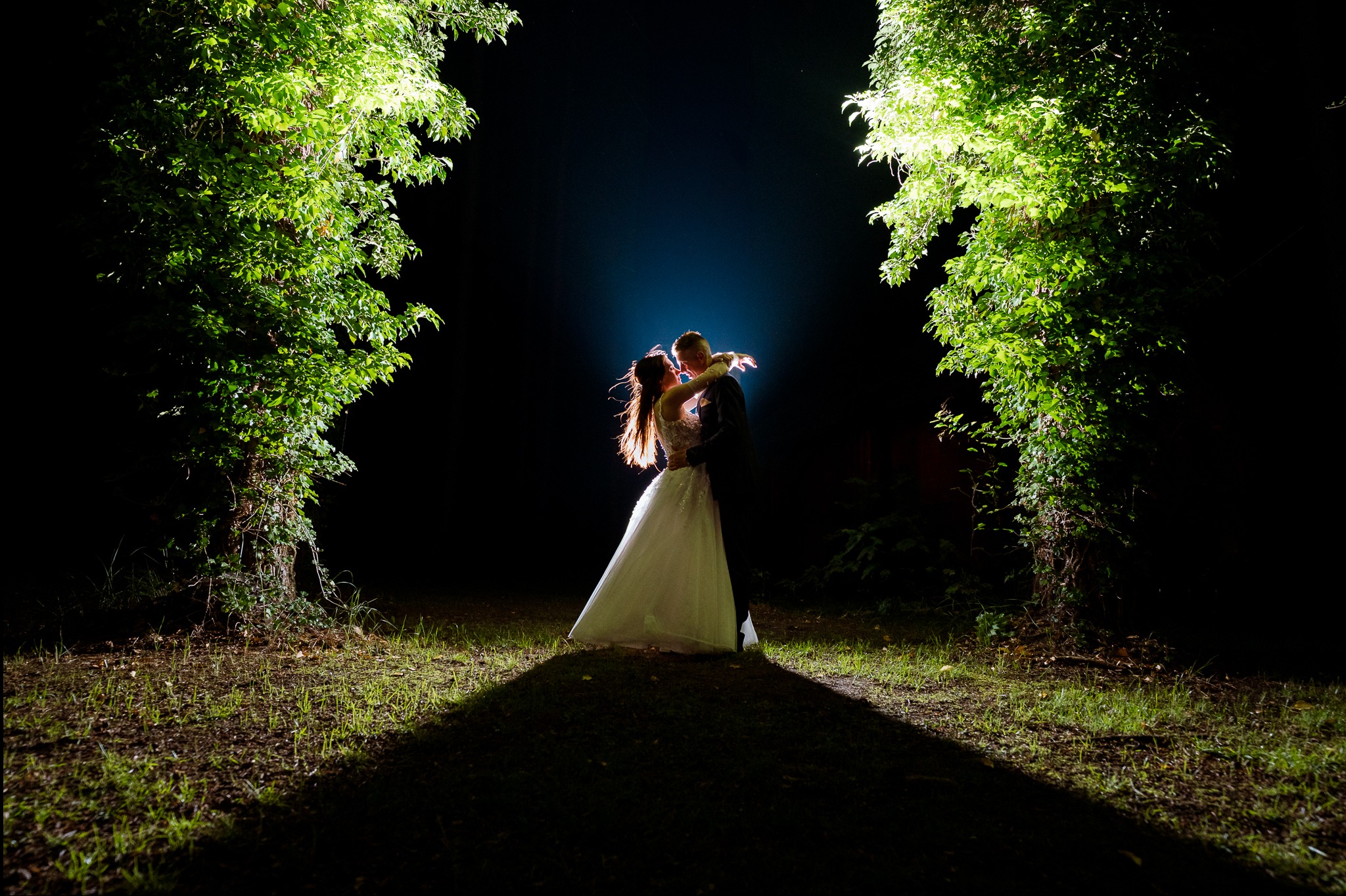 Para młoda w objęciach, nocą, oświetlona punktowo, otoczona drzewami. Romantyczna sceneria ślubna z unikalnym oświetleniem i kompozycją.
