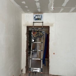 DMX Electrics - Rozdzielnia elektryczna zamontowana nad futryną, widoczna drabina z narzędziami. Ściany w trakcie wykończenia, surowy stan. Prace remontowe wewnątrz budynku.