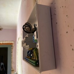 DMX Electrics - Otwarta skrzynka elektryczna z przewodami zamontowana na różowej ścianie w trakcie remontu. Widoczne elementy instalacji i przewody w kolorach niebieskim, żółtym i zielonym.