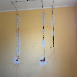 DMX Electrics - Żółta ściana z bruzdami wypełnionymi gipsem, w których poprowadzone są przewody elektryczne. Widoczne pomarańczowe puszki instalacyjne i kable przymocowane do sufitu.