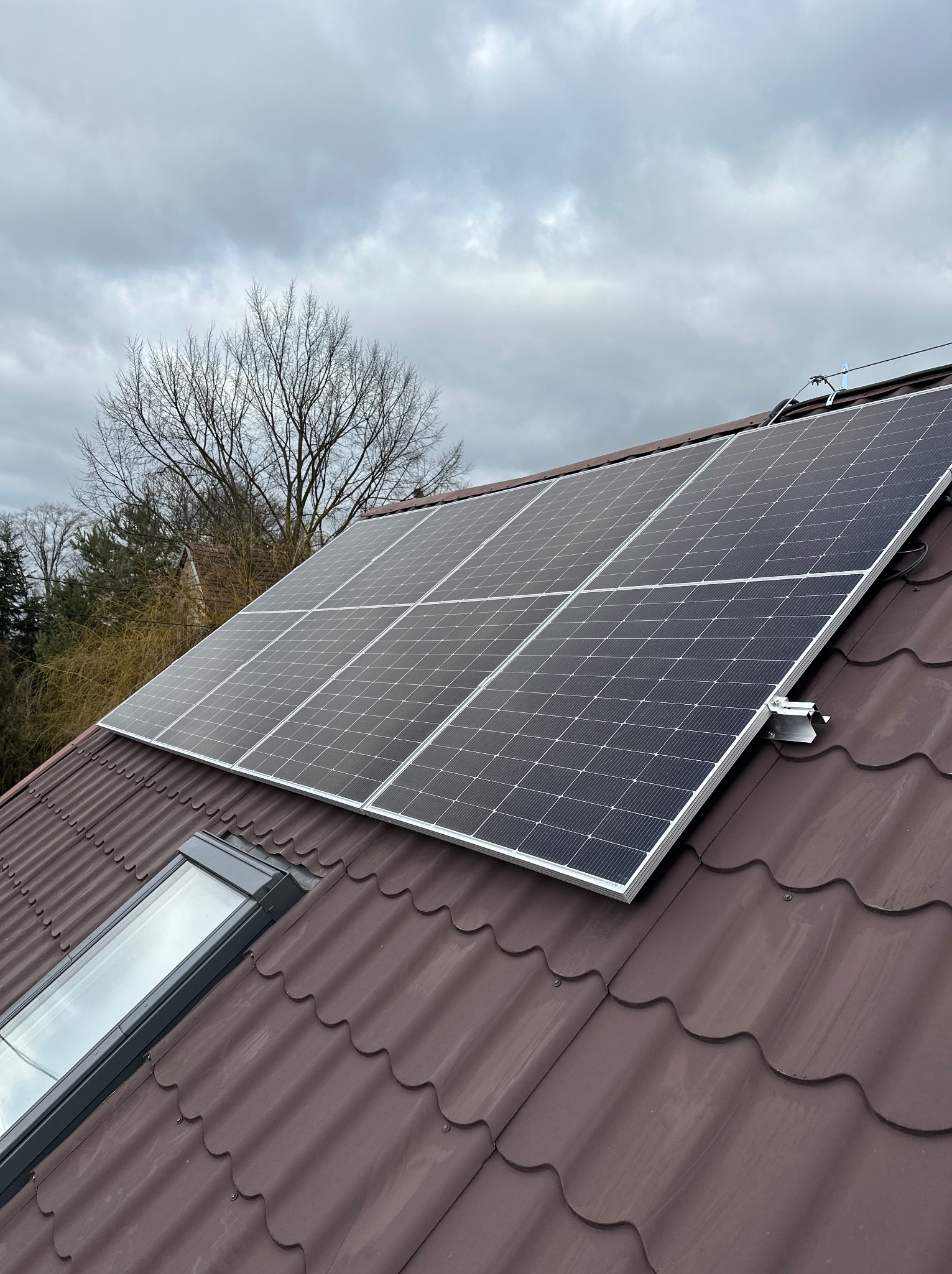 Panel słoneczny na brązowym dachu z dachówki, widok z góry, pochmurne niebo w tle. Widoczny fragment okna dachowego i drzewa bez liści.