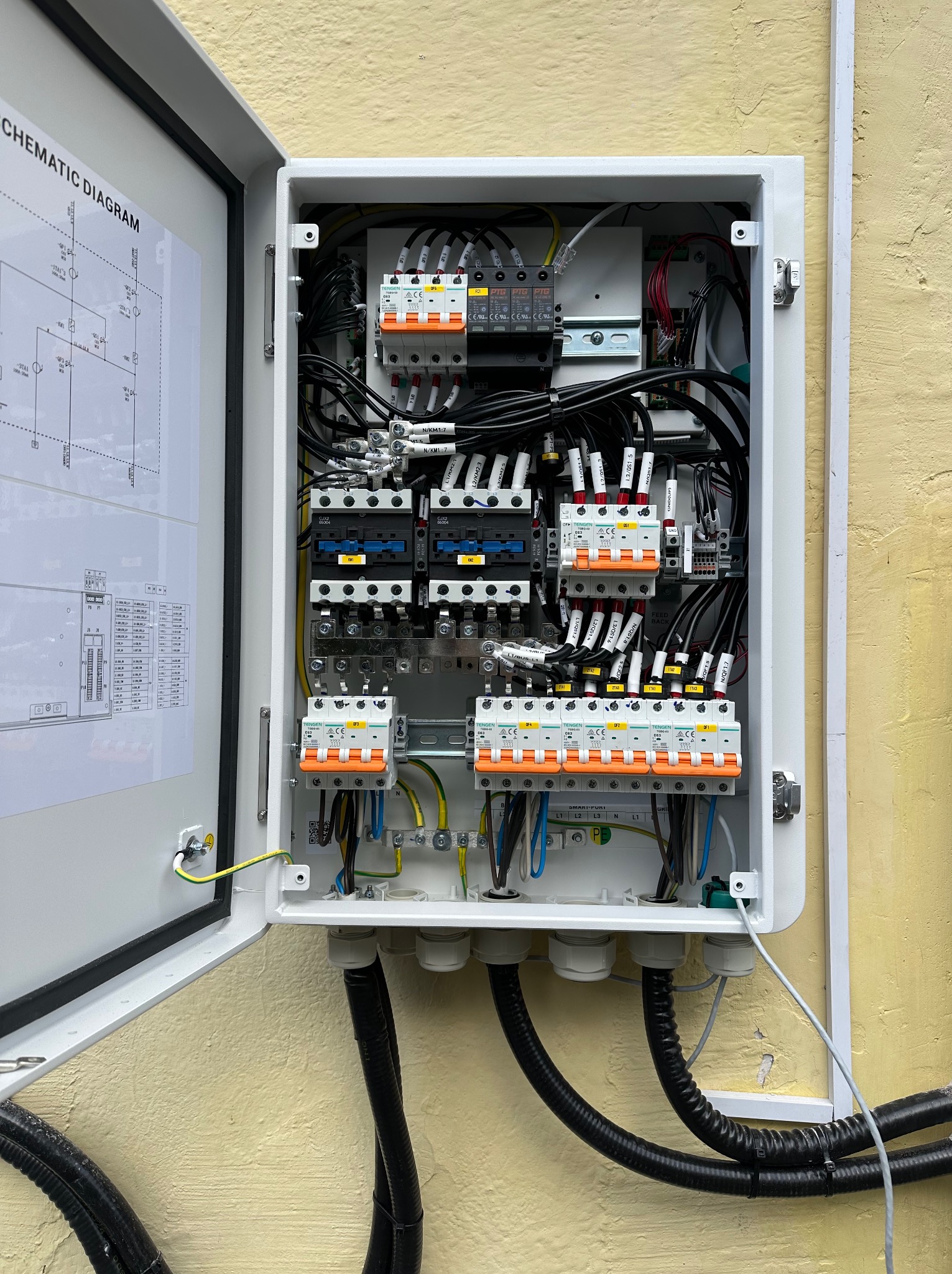 Otwarta skrzynka elektryczna z bezpiecznikami, przewodami i schematem na drzwiach, zamontowana na żółtej ścianie. Widoczne czarne kable wchodzące do skrzynki.