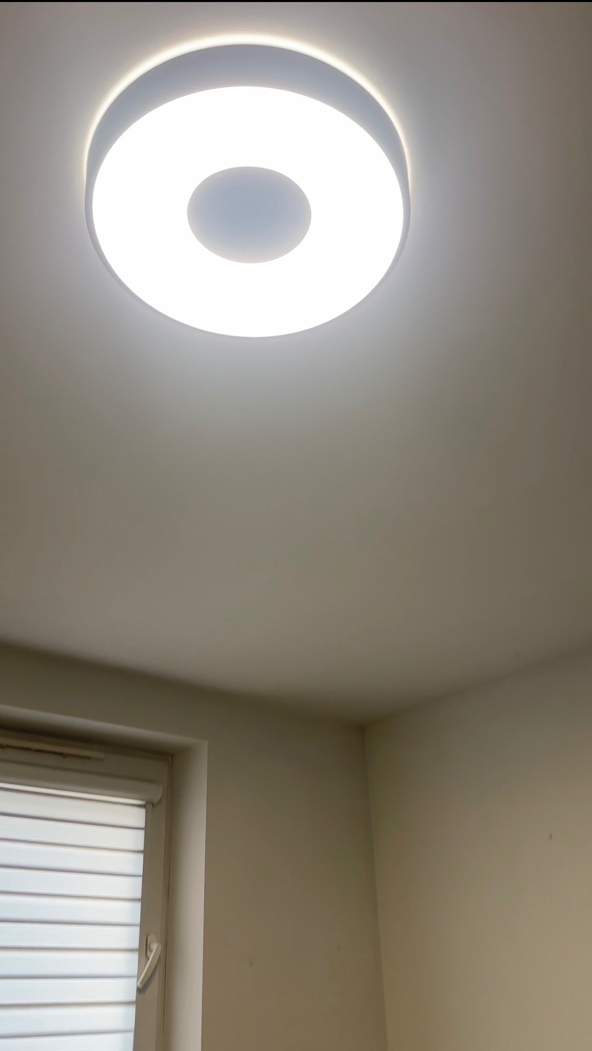 Zamontowana okrągła lampa sufitowa LED z białym światłem w pokoju z oknem z żaluzjami. Widok z dołu, neutralne tło ścian.