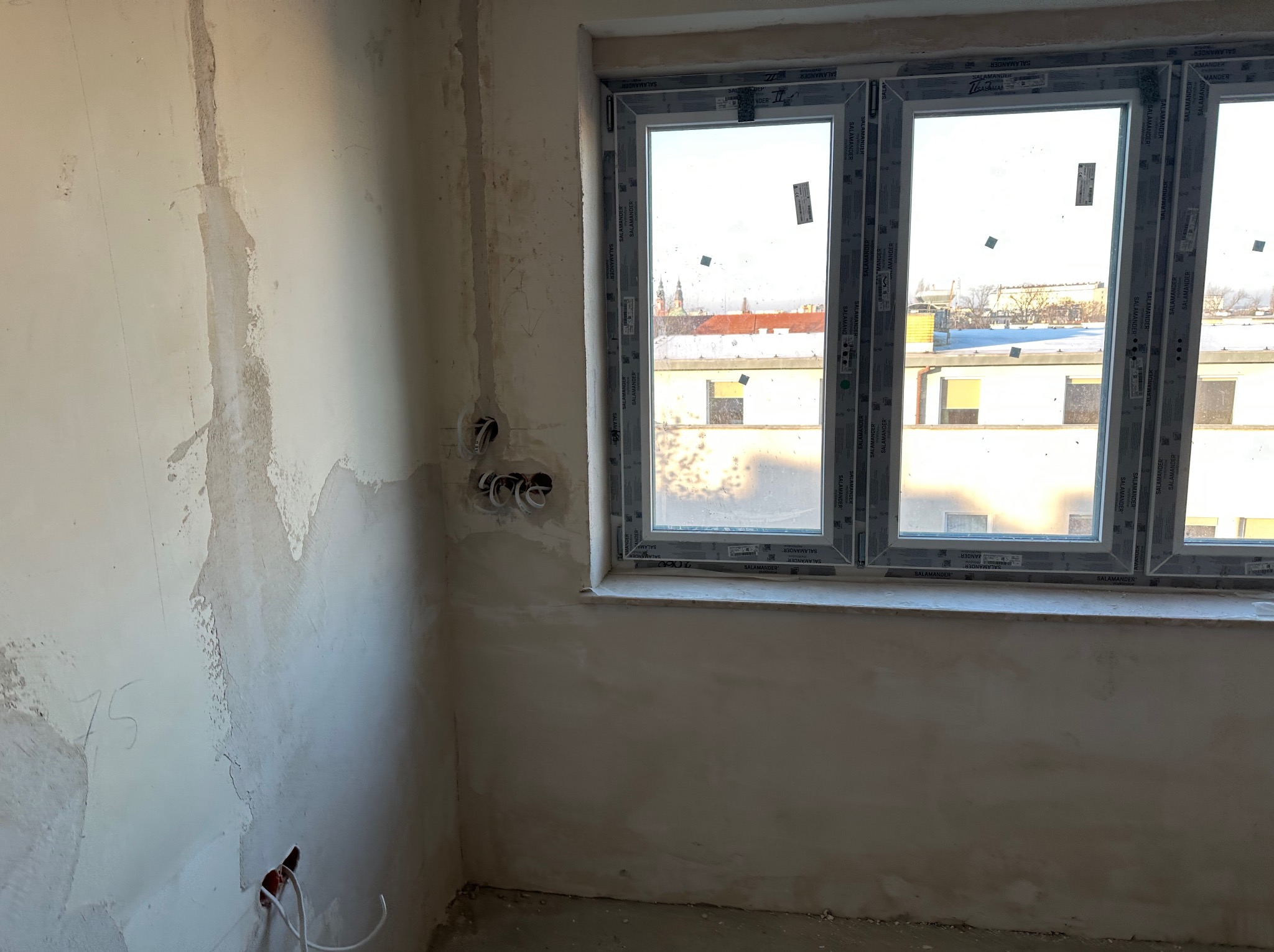 Surowy stan wnętrza: ściany po tynkowaniu z widocznymi puszkami elektrycznymi i wystającymi przewodami, okno z widokiem na miasto w tle.