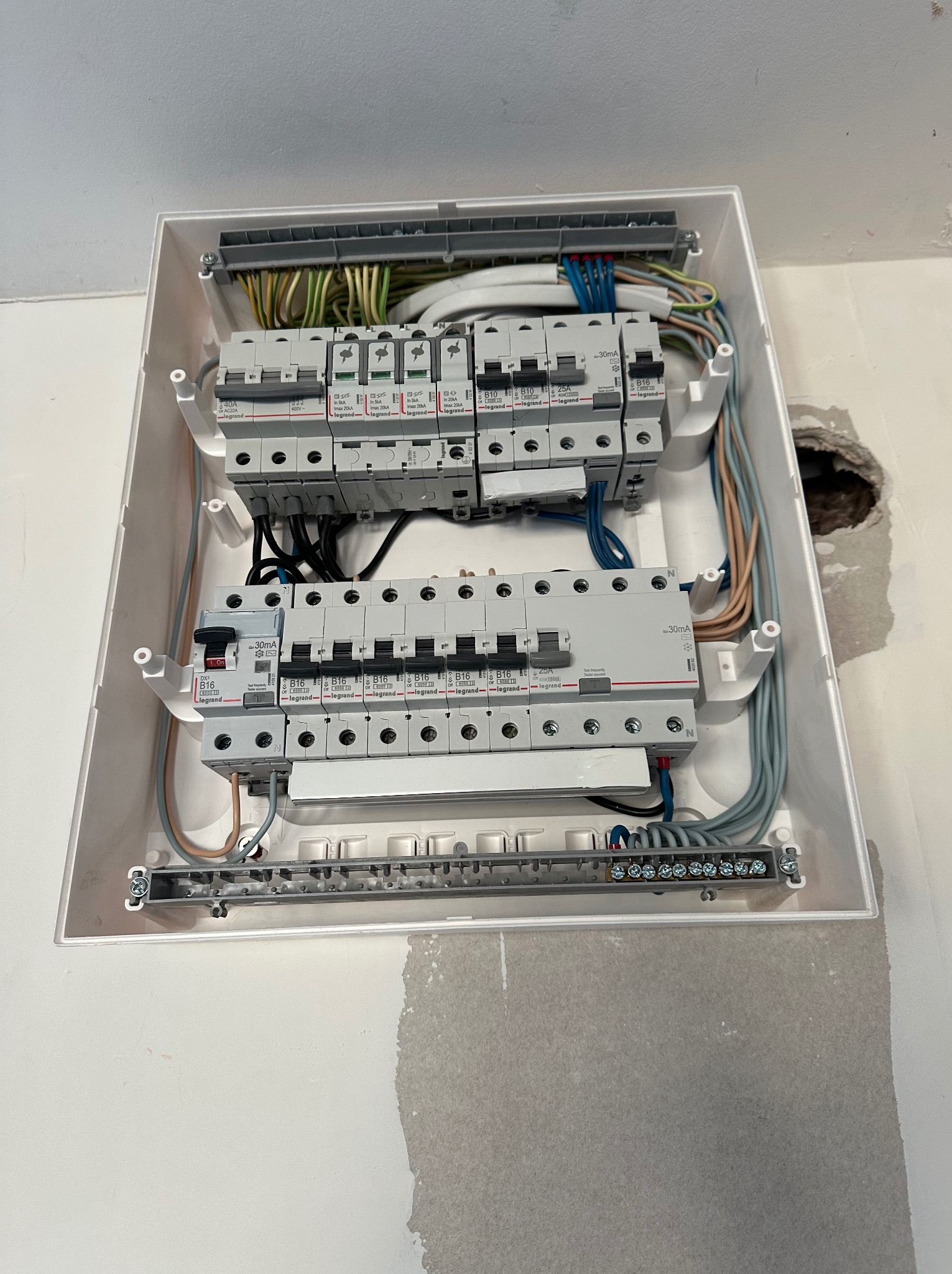 Rozdzielnica elektryczna w białej obudowie, widoczne bezpieczniki i przewody. Ściana wokół w stanie surowym, z widocznymi śladami po pracach instalacyjnych.