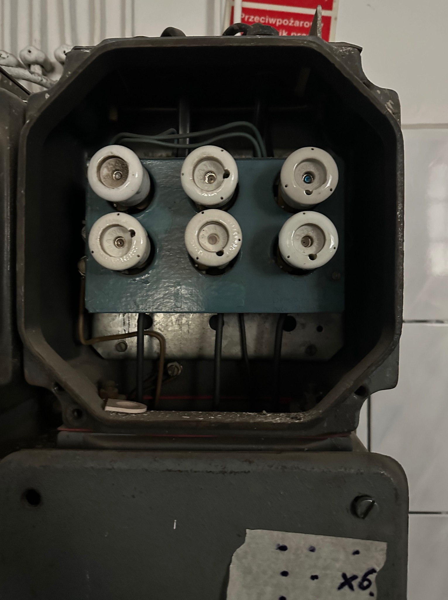 Otwarta skrzynka bezpiecznikowa z sześcioma ceramicznymi bezpiecznikami topikowymi na niebieskiej płycie montażowej. Widoczne przewody i elementy konstrukcyjne.