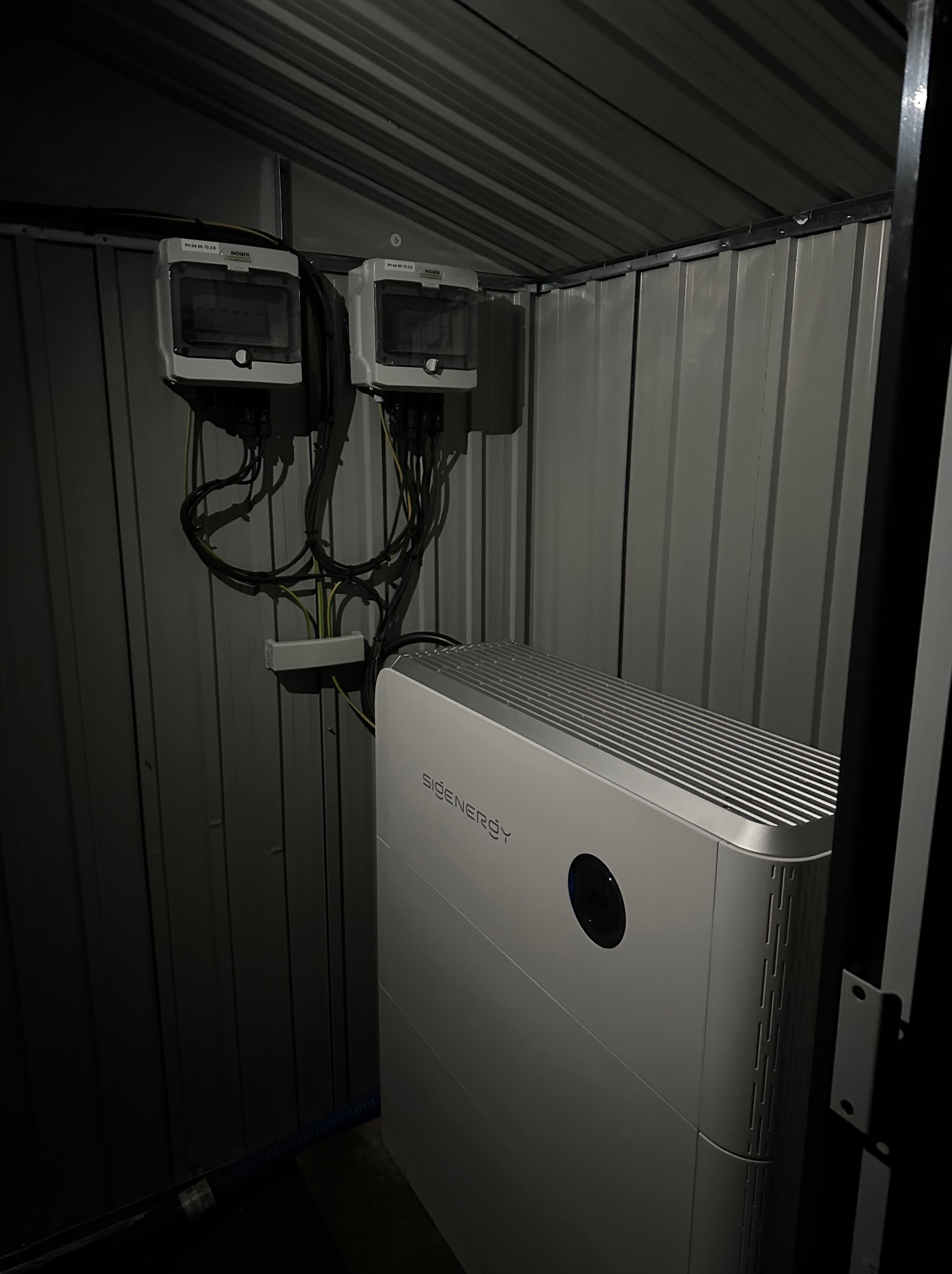 Wnętrze metalowego pomieszczenia z zamontowanymi dwoma licznikami NOARK i akumulatorem Sidenery. Widoczne kable i przewody elektryczne. Słabe oświetlenie.