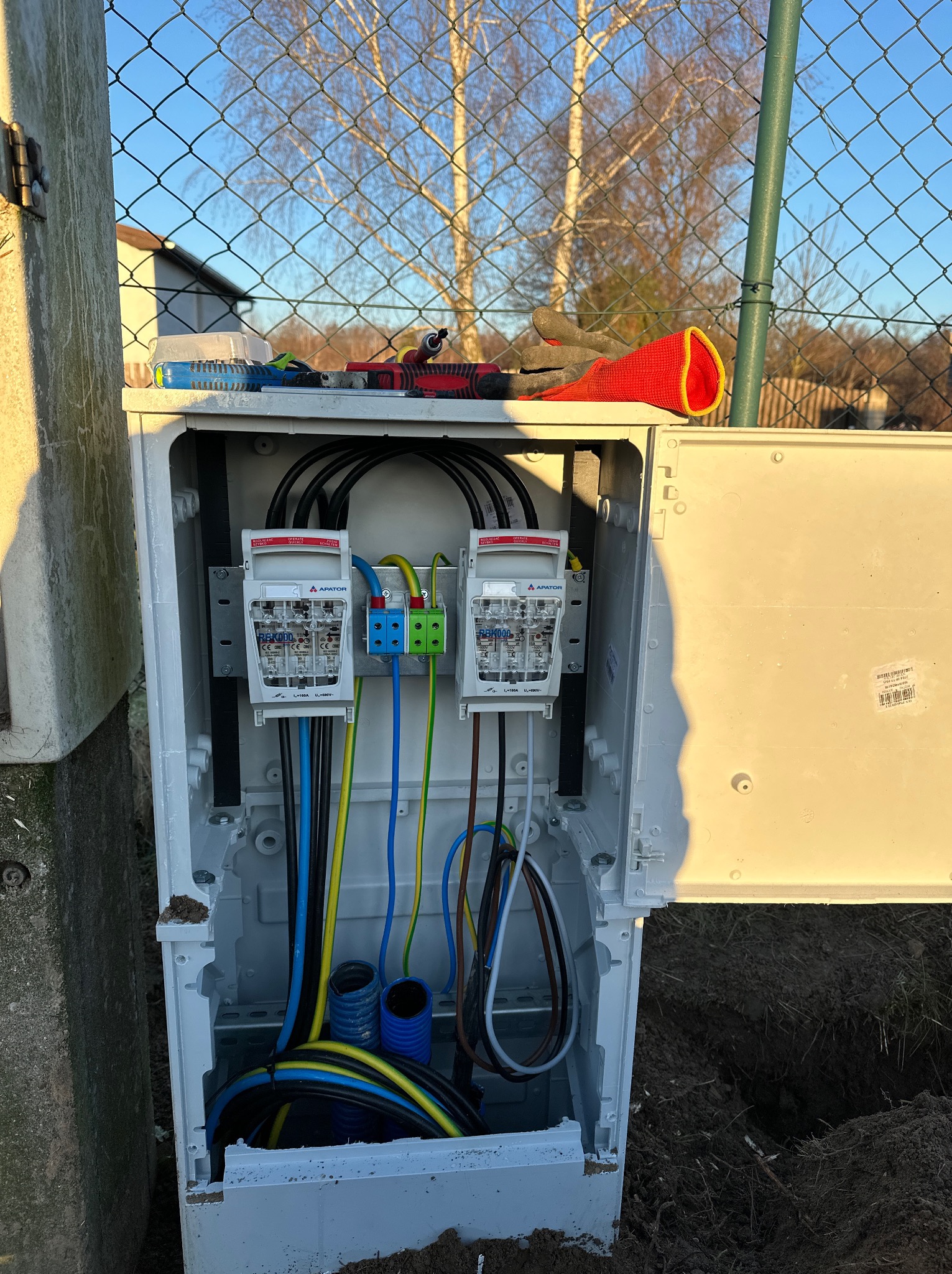 Otwarta skrzynka z dwoma licznikami energii elektrycznej, widoczne kable w kolorach: niebieskim, żółtym, czarnym i brązowym. Na górze skrzynki leżą narzędzia.