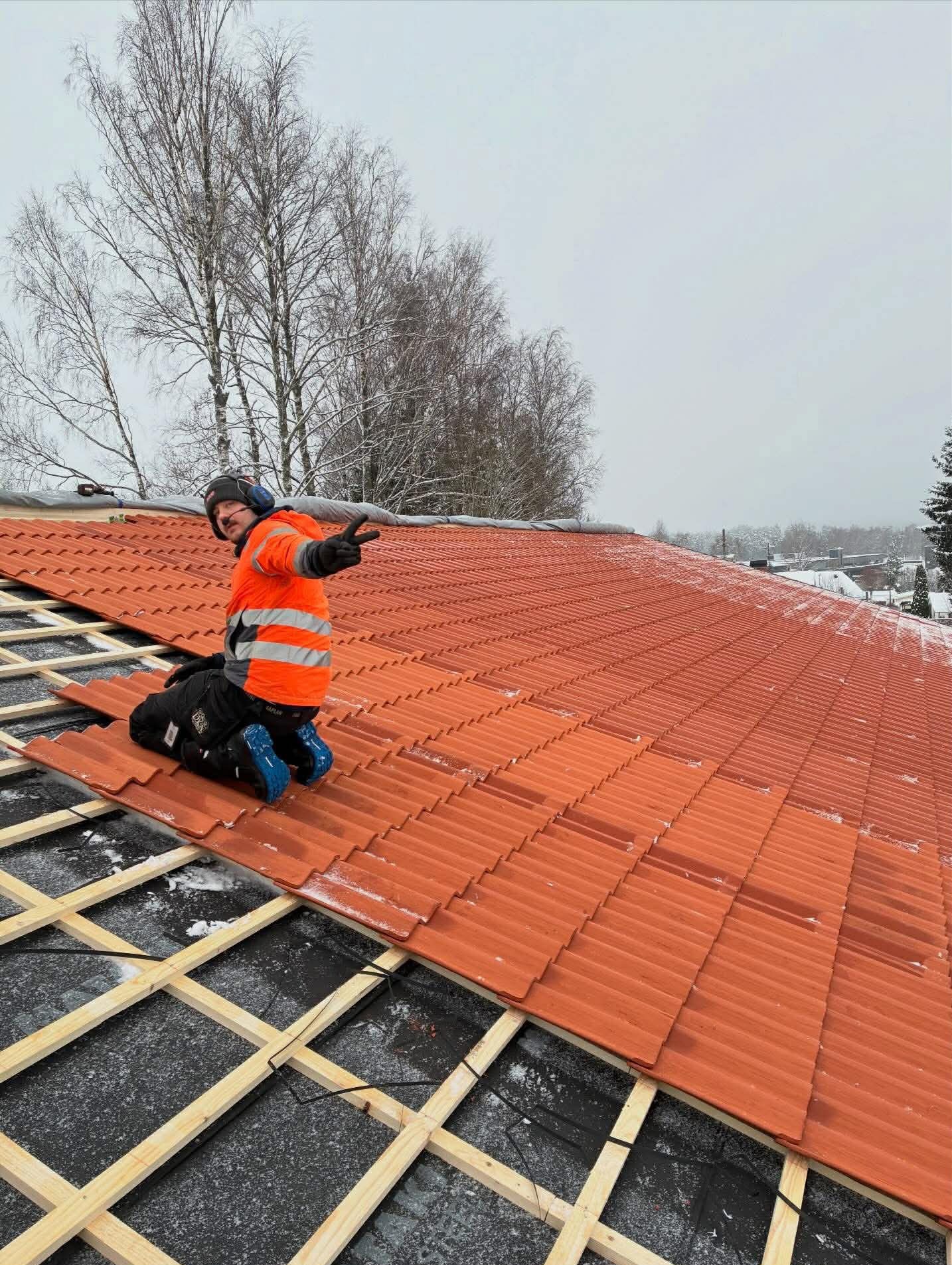 Dekarz w pomarańczowej kurtce klęczy na nowym, pomarańczowym dachu z dachówki ceramicznej, wskazując palcem. W tle drzewa i pochmurne niebo.