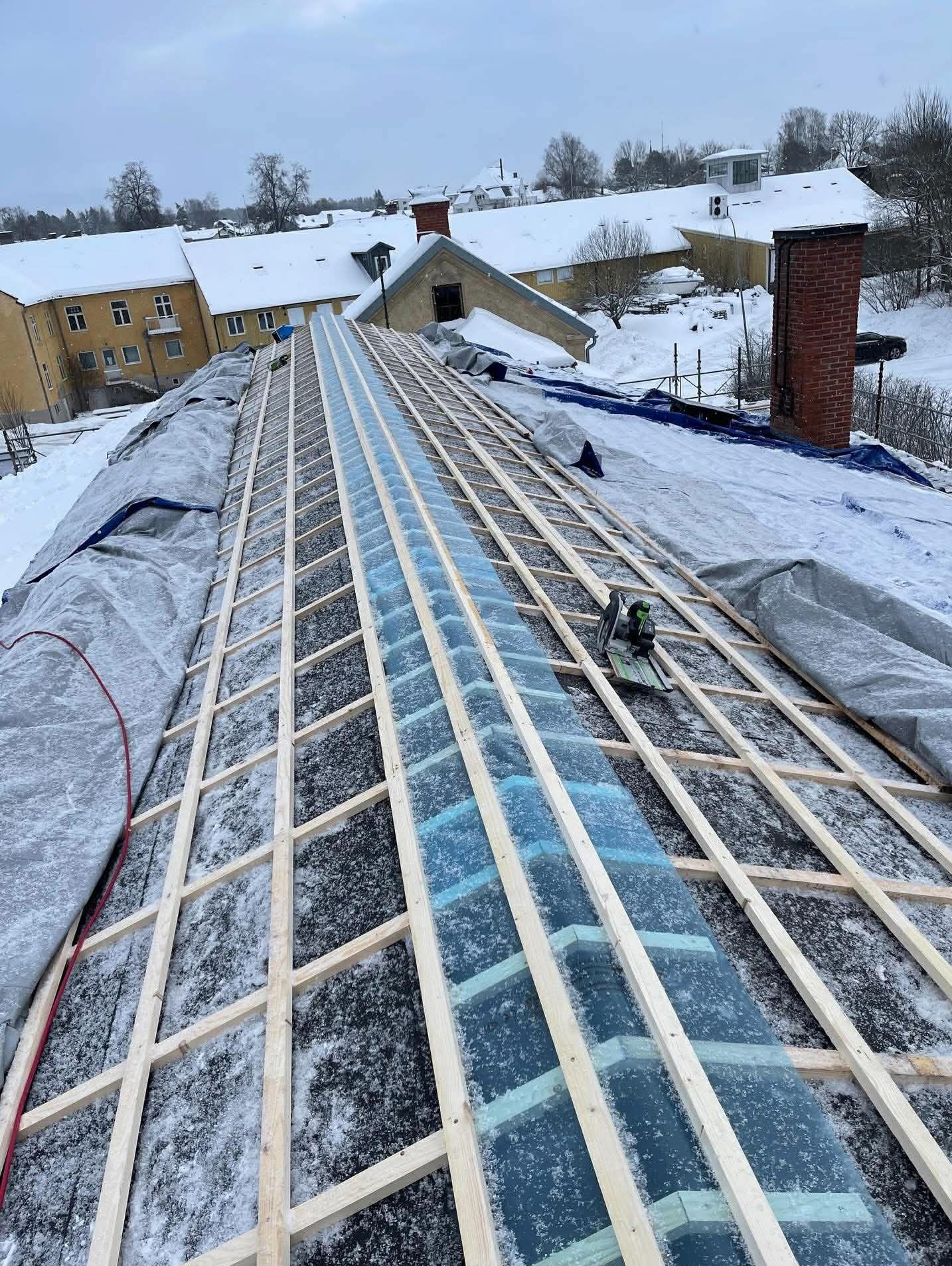 W trakcie prac na dachu: drewniana konstrukcja, częściowo przykryta folią, z widoczną deską i narzędziem. Zimowy krajobraz w tle.