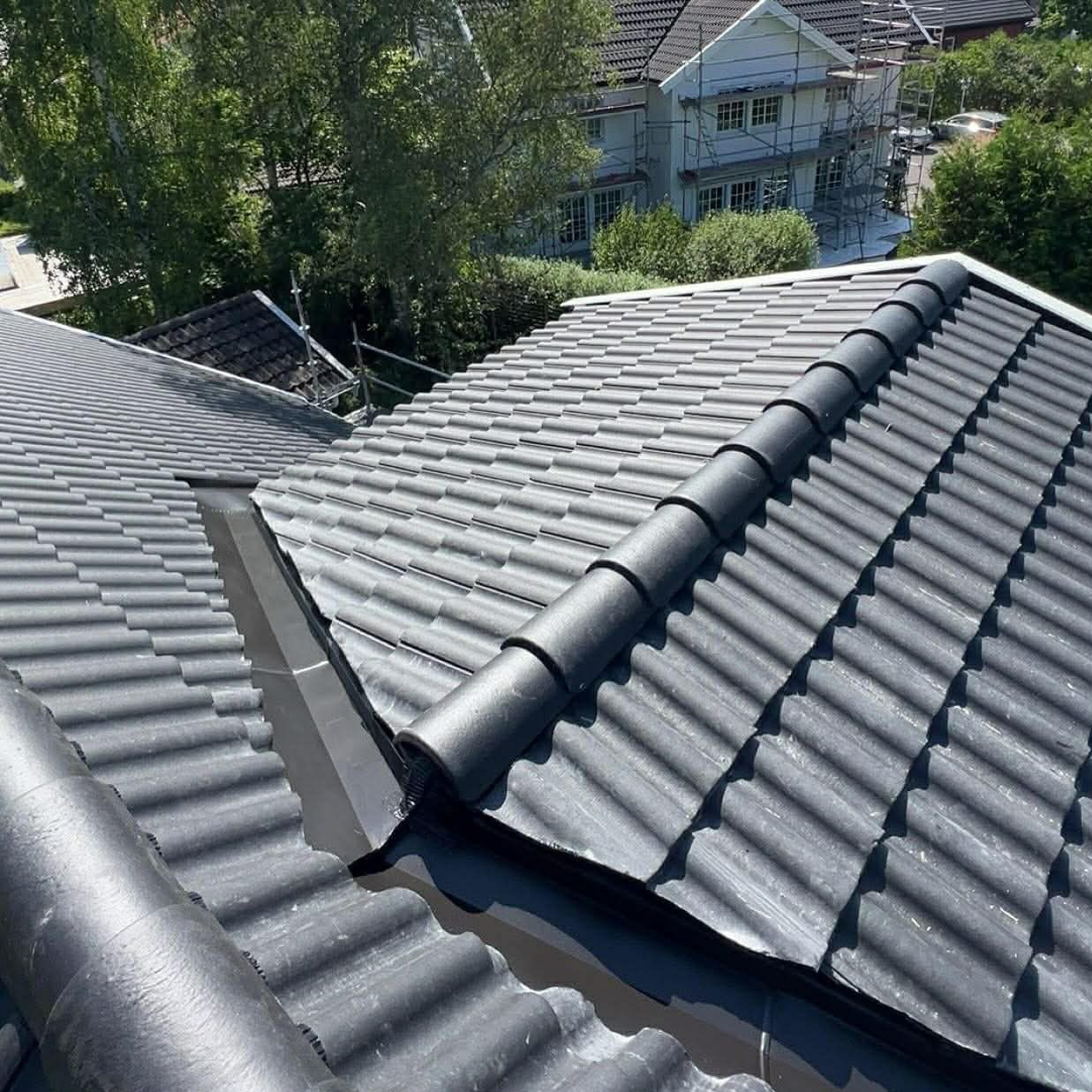 Ciemnoszary dach z ceramicznej dachówki karpiówki, widok z góry. W tle dom w rusztowaniach, drzewa i błękitne niebo. Układanie dachówki w rzędach.