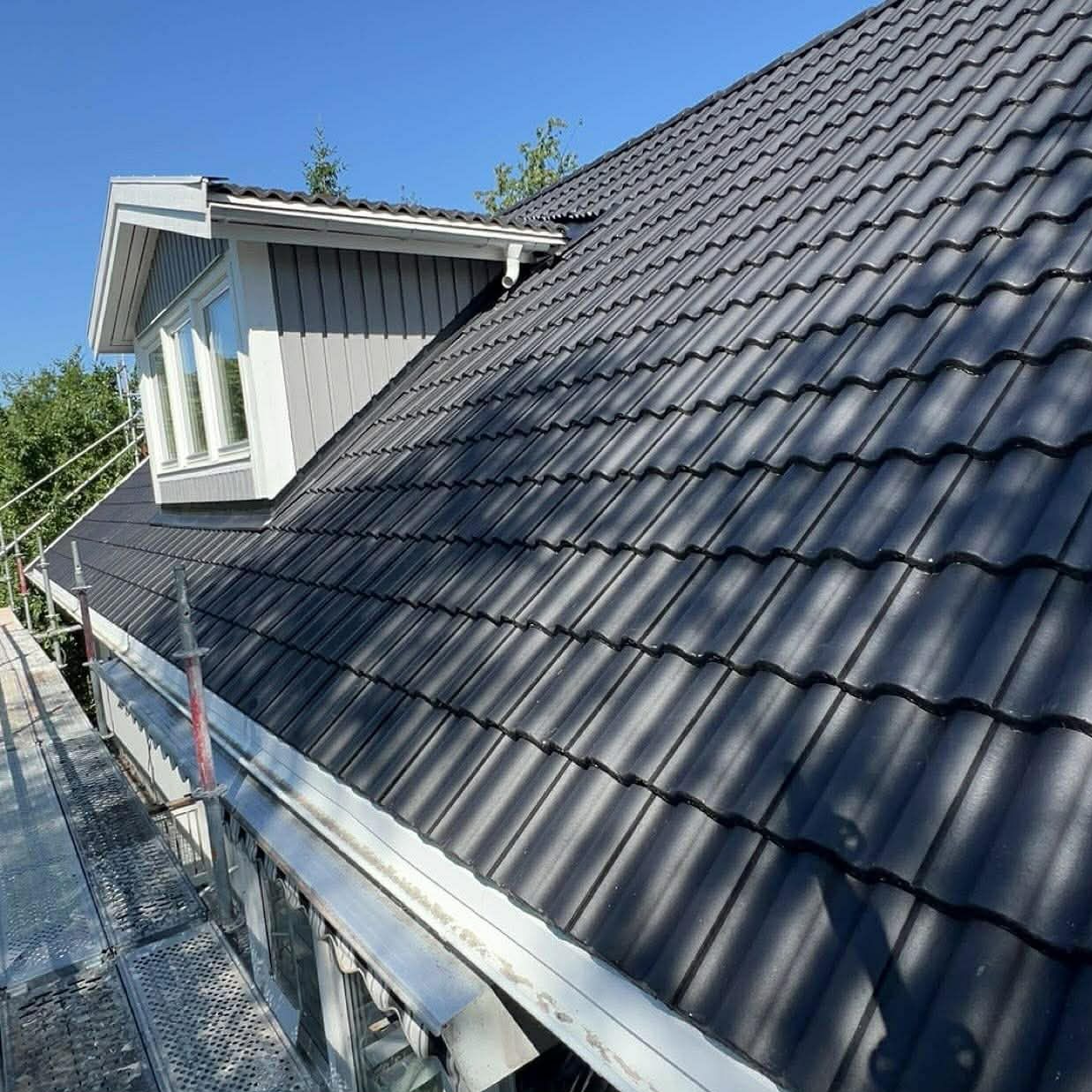 Ciemna dachówka ceramiczna na skośnym dachu z lukarną, widok z rusztowania. Rynna w kolorze srebrnym. Błękitne niebo w tle.