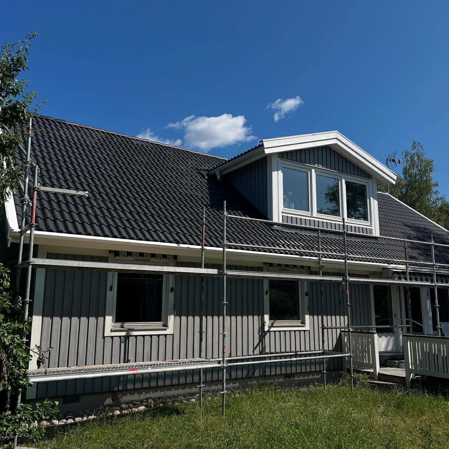 Dom jednorodzinny z szarą elewacją i ciemnym dachem, otoczony rusztowaniem. Widoczne okna dachowe i fragment zielonego trawnika. Błękitne niebo z pojedynczymi chmurami.