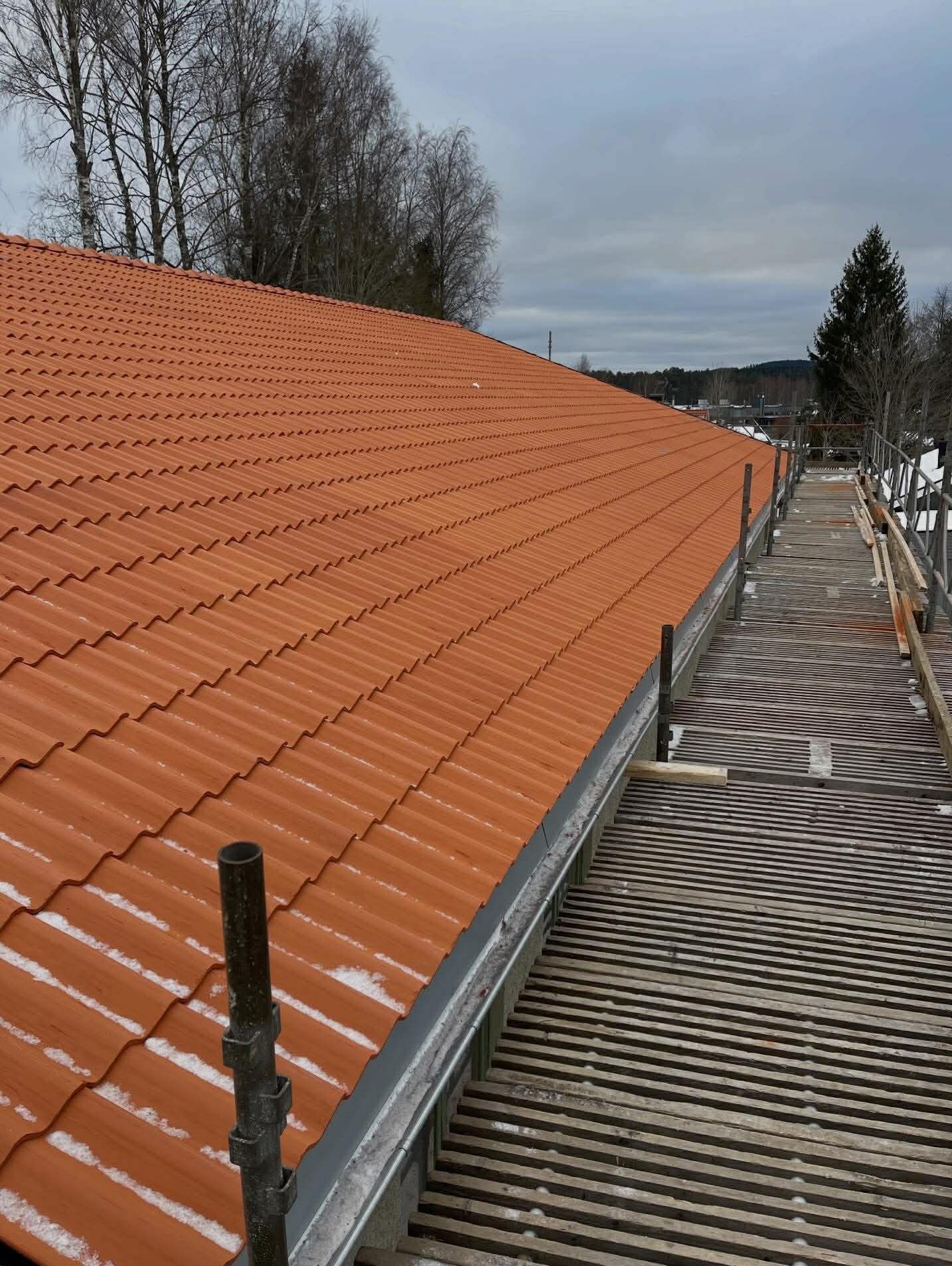 Czerwona dachówka ceramiczna na skośnym dachu budynku, widok z góry, z fragmentem rusztowania i zimowym krajobrazem w tle. Widoczne ślady śniegu.