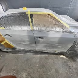 Rostyslav Tomilenko - Auto przygotowane do lakierowania: oklejone folią i taśmą, widoczne drzwi i błotnik w szarym podkładzie, oświetlenie warsztatowe.