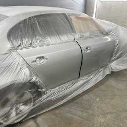 Rostyslav Tomilenko - Samochód osobowy w trakcie lakierowania, zabezpieczony folią ochronną, widoczne świeżo polakierowane drzwi w odcieniach szarości, detale karoserii zaklejone taśmą.