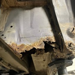 AutoRevive naprawy lakiernicze-blacharskie - Silnie skorodowany fragment podwozia samochodu na podnośniku, widoczne dziury i rdza. Ujęcie z dołu, oświetlenie warsztatowe.