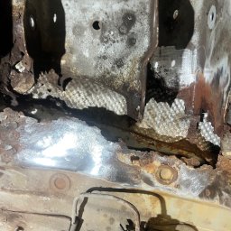 AutoRevive naprawy lakiernicze-blacharskie - Zbliżenie na skorodowaną podłogę samochodu w trakcie naprawy. Widoczne ogniska rdzy i fragmenty oczyszczone do gołej blachy. Elementy zawieszenia w dolnej części kadru.