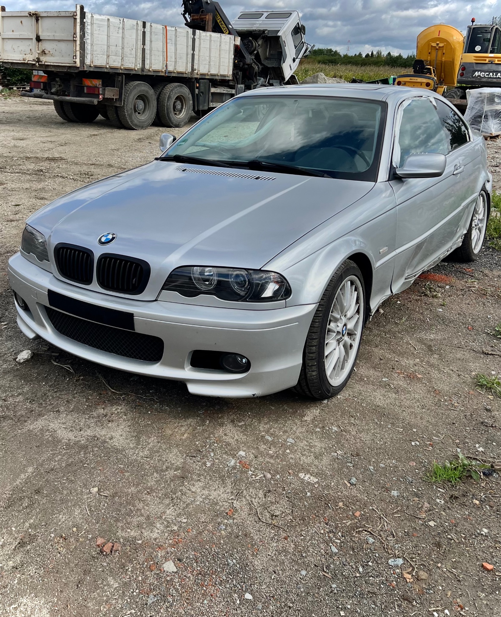 Srebrne BMW E46 Coupe z uszkodzeniami karoserii, zaparkowane na placu budowy. Widoczne wgniecenia na progu i błotniku. W tle ciężarówka i maszyny budowlane.