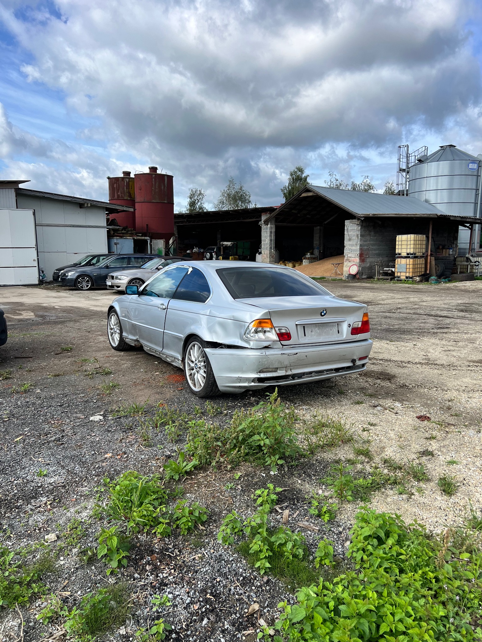 Srebrne BMW E46 Coupe z uszkodzeniami karoserii, widoczne wgniecenia i brakujące elementy. Tło stanowią budynki gospodarcze i pochmurne niebo.