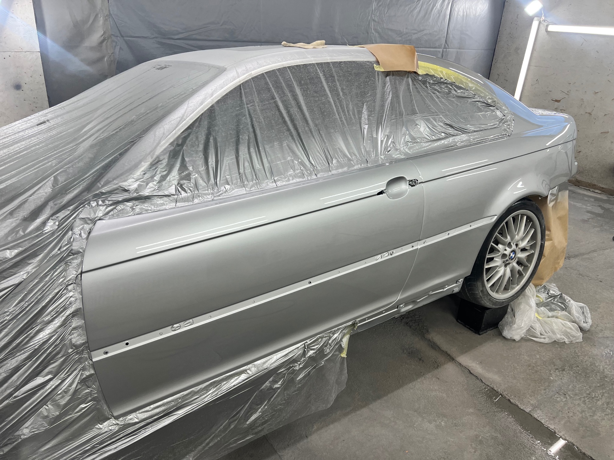Srebrny samochód BMW serii 3 E46, częściowo zakryty folią ochronną i taśmą malarską, przygotowany do lakierowania w warsztacie. Widoczne koło i fragment karoserii.