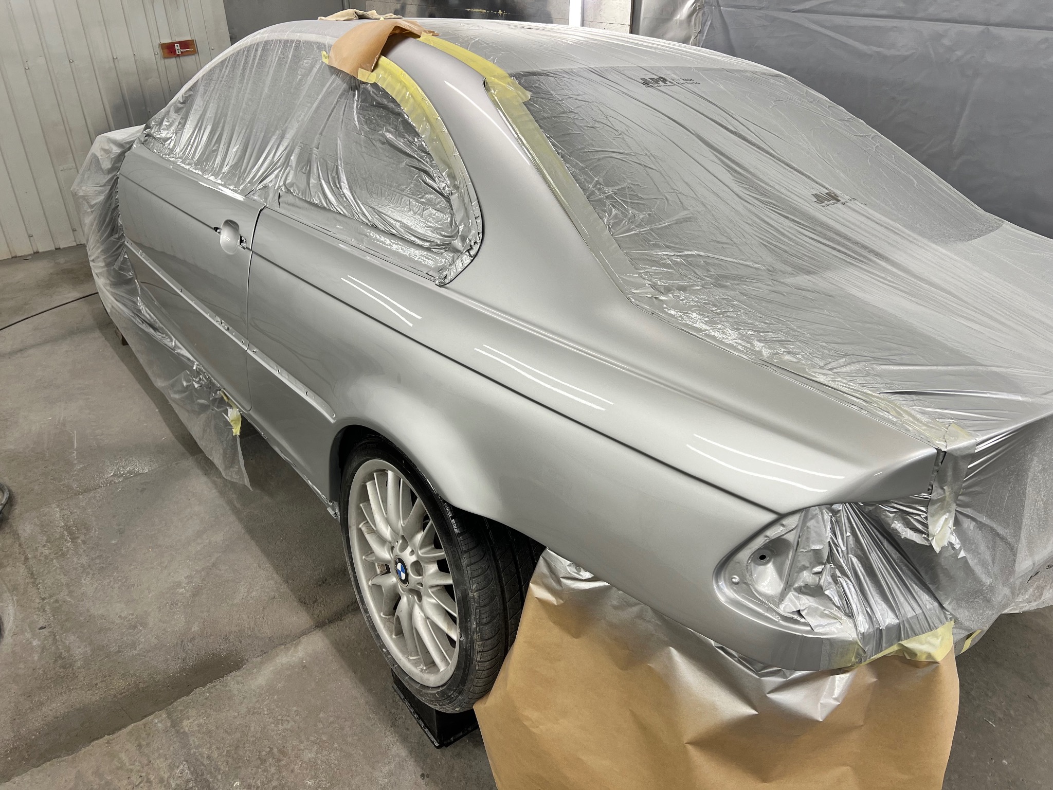 Srebrne BMW serii 3 E46 coupe, przygotowane do lakierowania, zabezpieczone folią i taśmą maskującą w warsztacie. Widoczny fragment koła i opony.