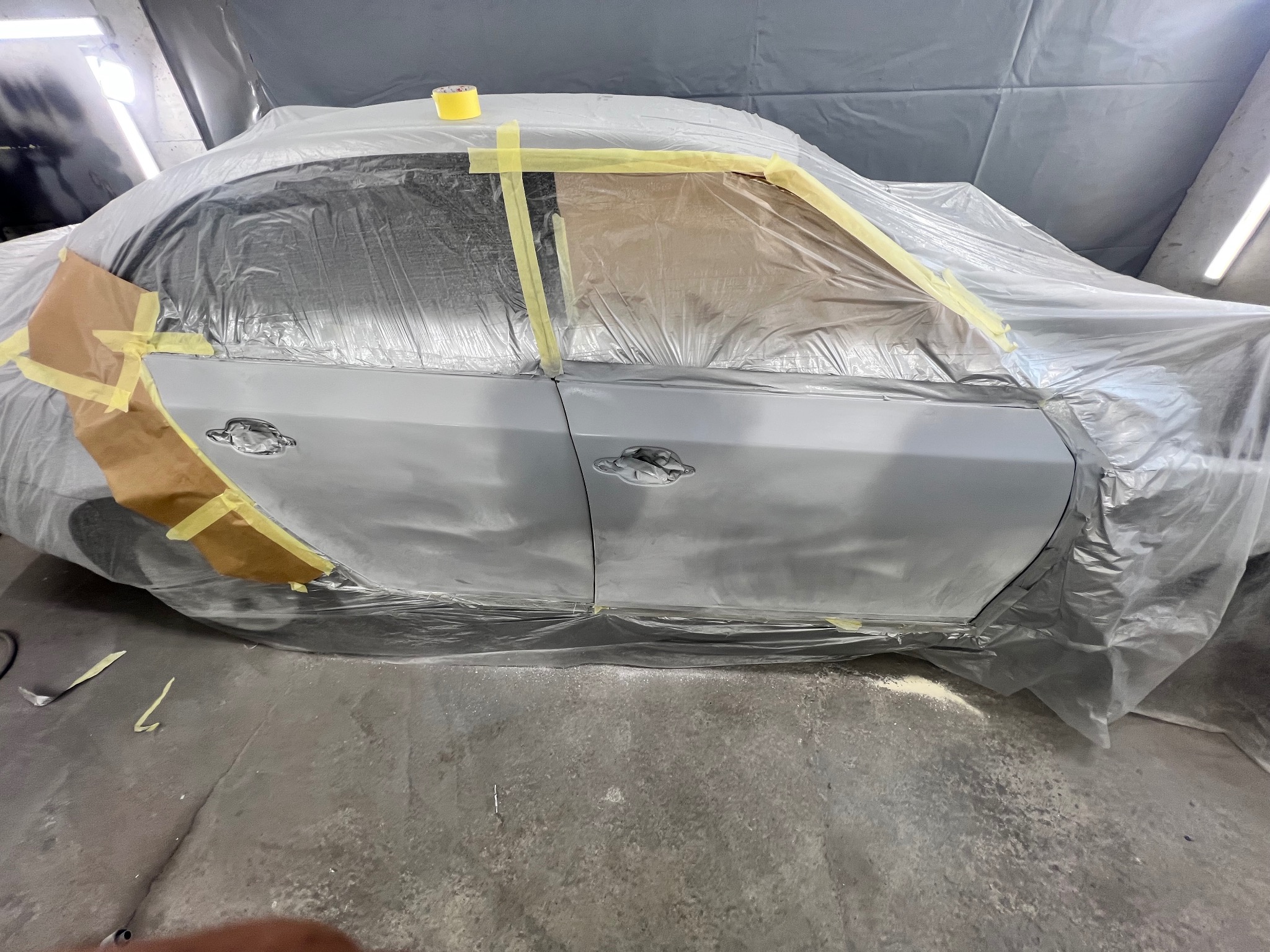 Auto przygotowane do lakierowania: oklejone folią i taśmą, widoczne drzwi i błotnik w szarym podkładzie, oświetlenie warsztatowe.