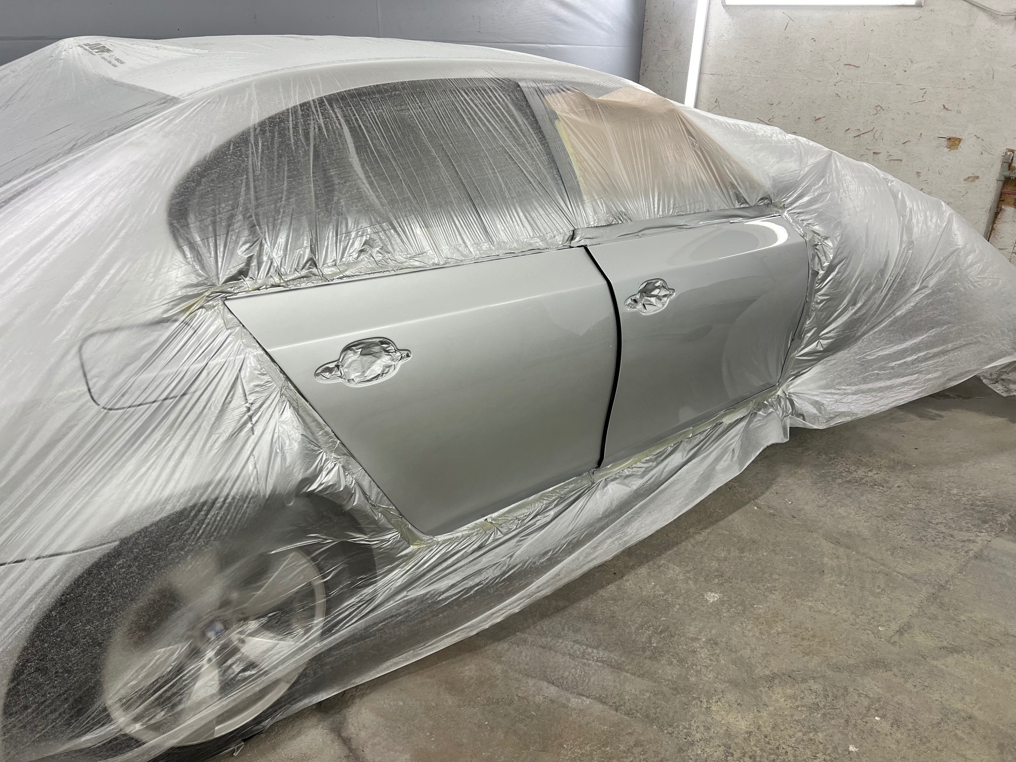 Samochód osobowy w trakcie lakierowania, zabezpieczony folią ochronną, widoczne świeżo polakierowane drzwi w odcieniach szarości, detale karoserii zaklejone taśmą.