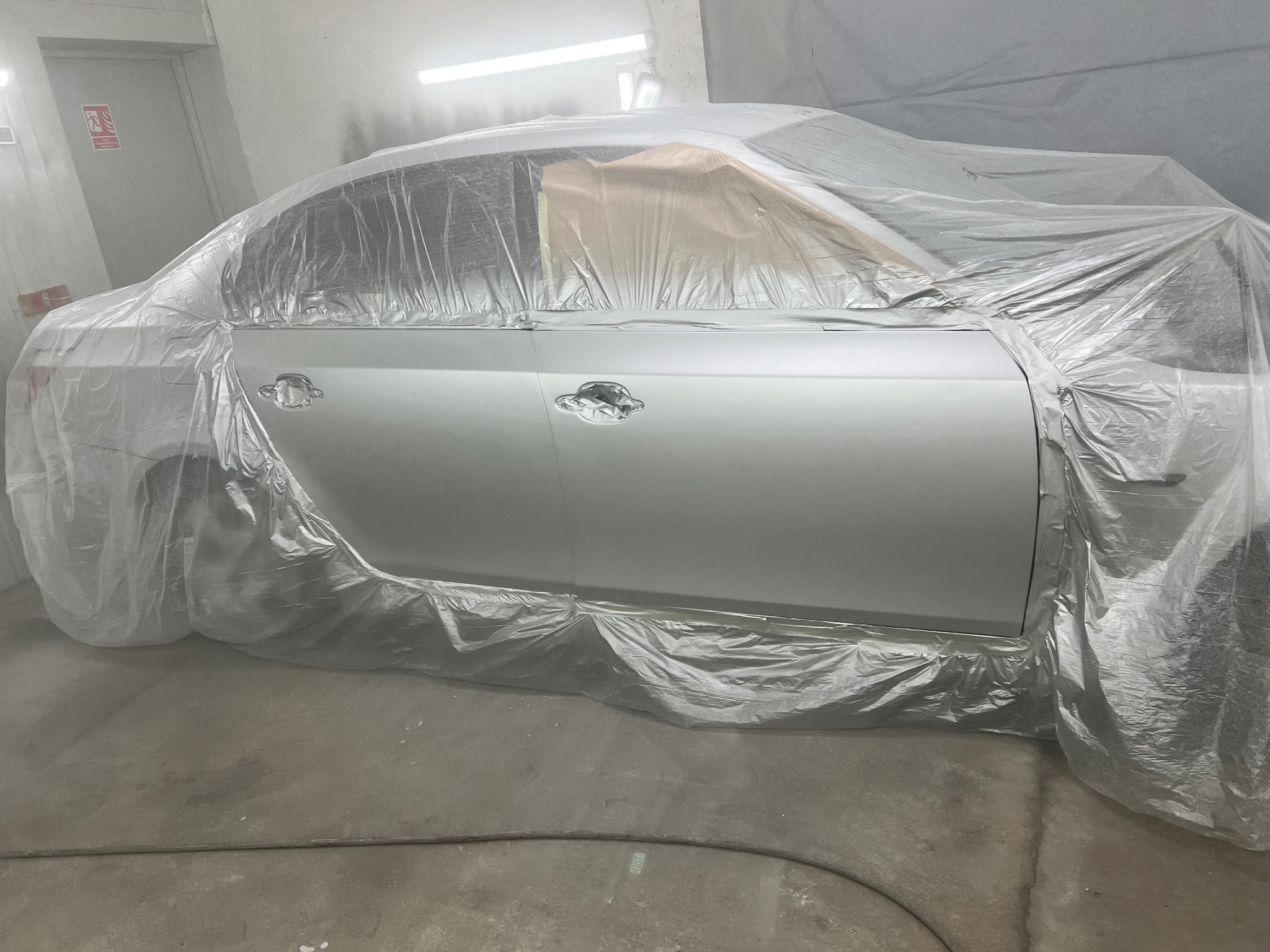 Samochód osobowy w komorze lakierniczej, zabezpieczony folią przed lakierowaniem. Widoczne detale karoserii i fragment podłogi warsztatu. Oświetlenie górne.