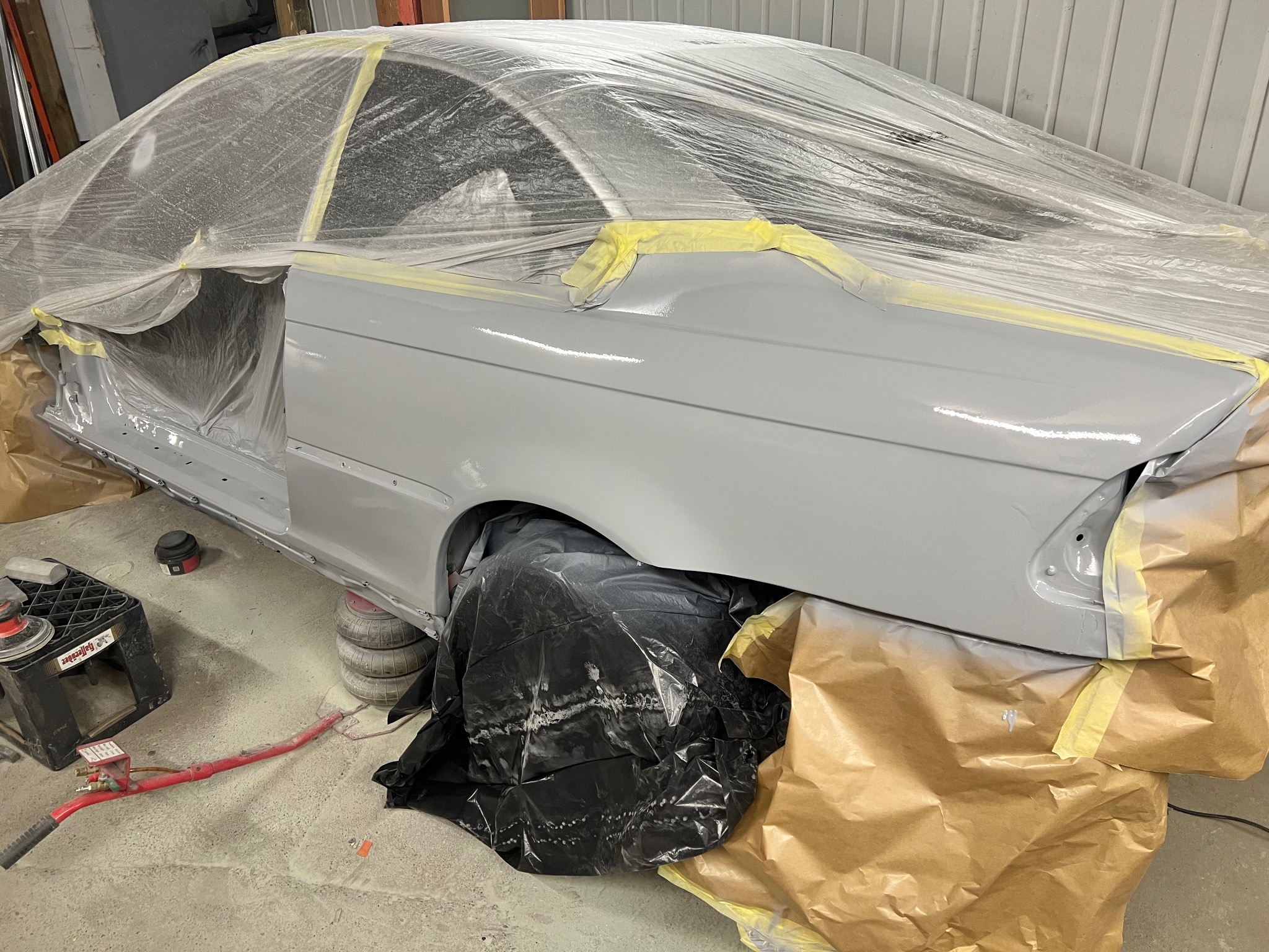 Szary samochód w trakcie lakierowania, zabezpieczony folią i taśmą malarską. Widoczne narzędzia i przygotowane podłoże. Detal z warsztatu blacharsko-lakierniczego.