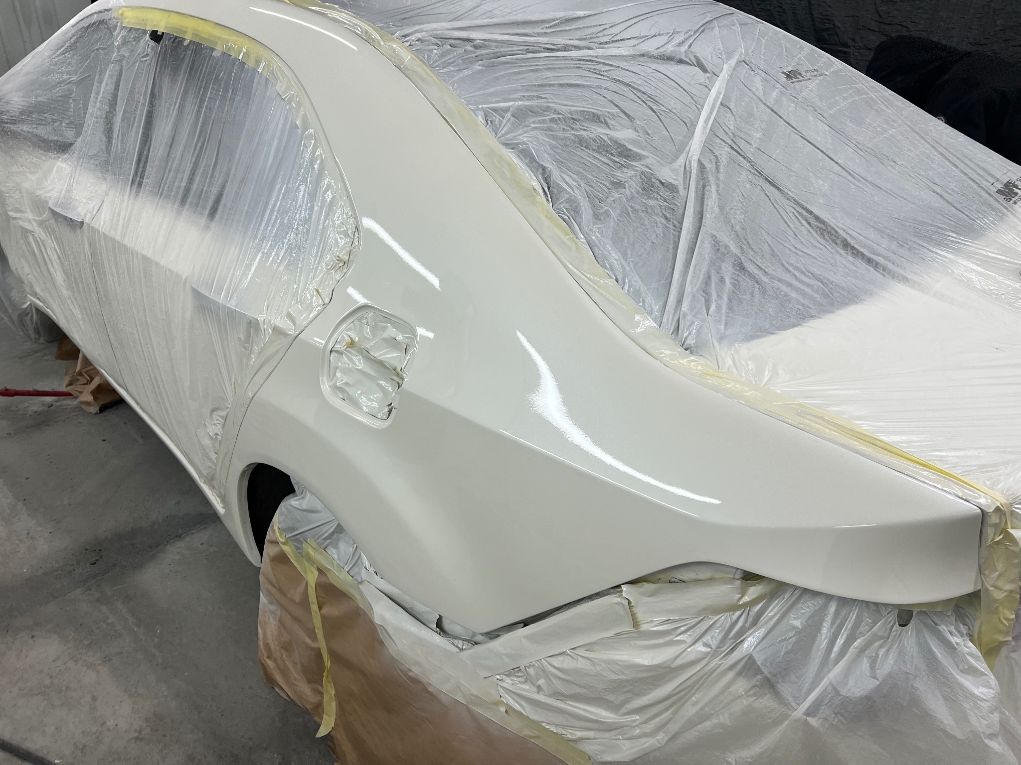 Biały samochód w trakcie lakierowania, zabezpieczony folią i taśmą malarską. Widoczny fragment karoserii po nałożeniu świeżej warstwy lakieru.