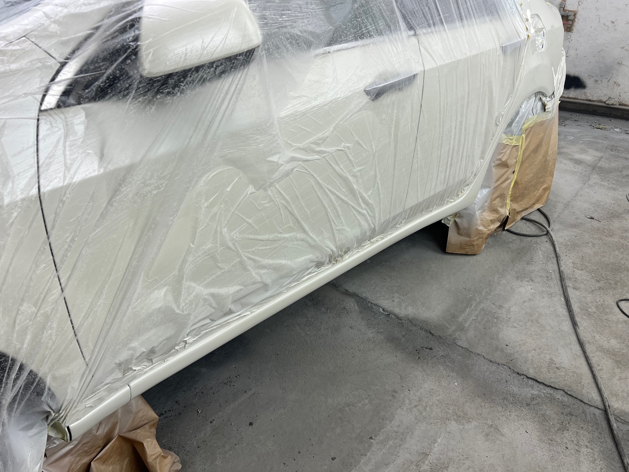 Biały samochód w folii ochronnej i papierze maskującym, przygotowany do lakierowania w warsztacie. Widoczne drzwi, błotnik i próg. Szary beton na podłodze.