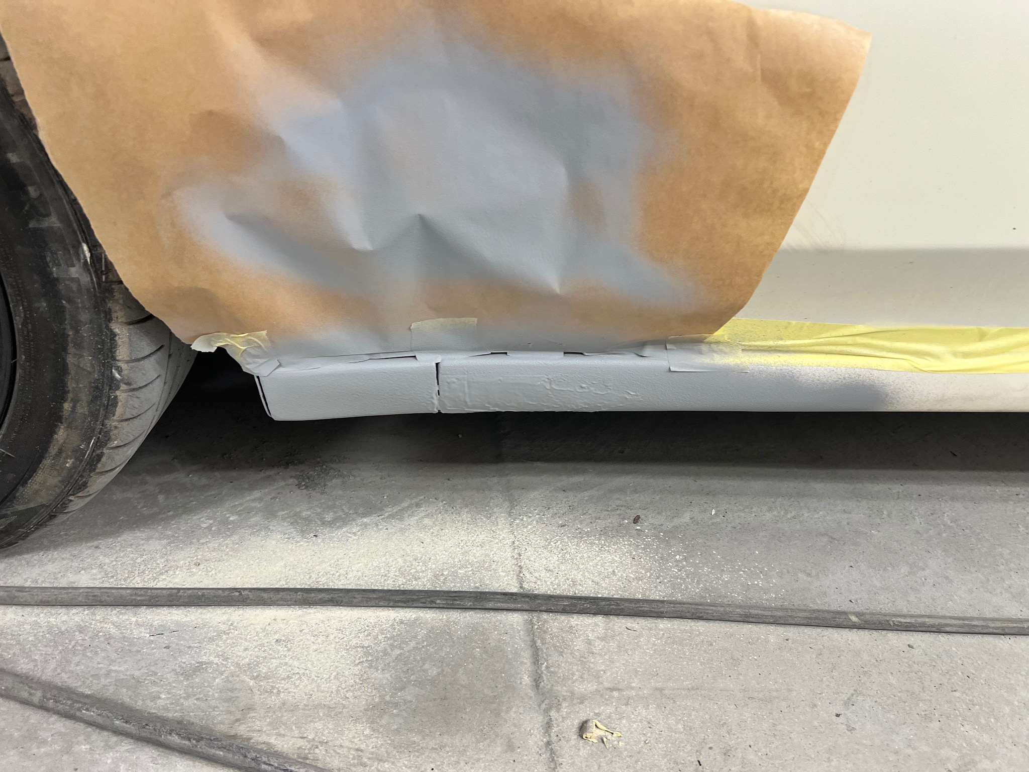 Fragment karoserii auta w trakcie lakierowania: zabezpieczony papierem i taśmą fragment nadkola, świeżo pomalowany próg w szarym kolorze. Widoczne koło i podłoga warsztatu.