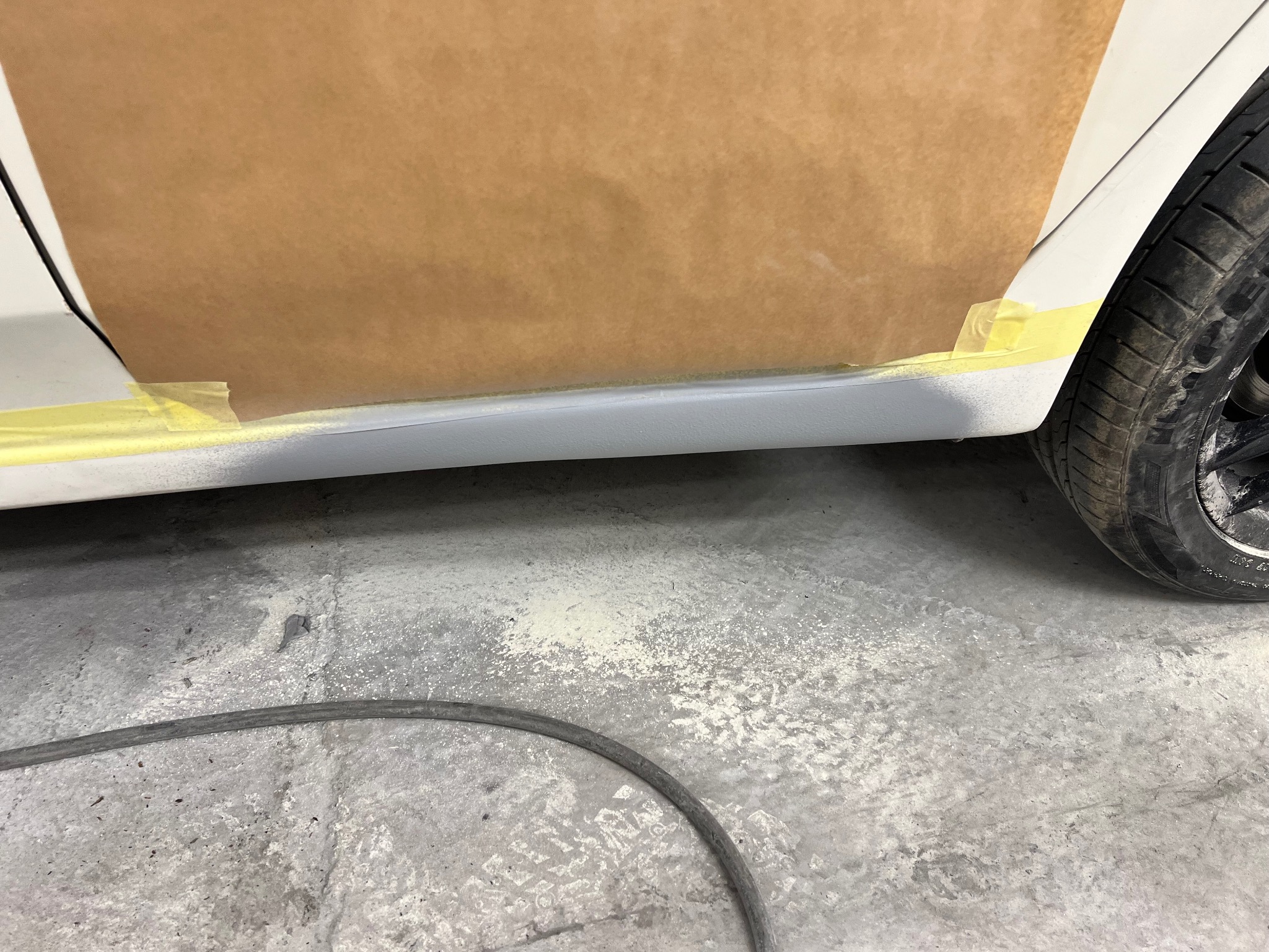 Przygotowanie do lakierowania: fragment karoserii auta z maskowaniem papierem i taśmą. Widoczny fragment opony i przewód na podłodze warsztatu.