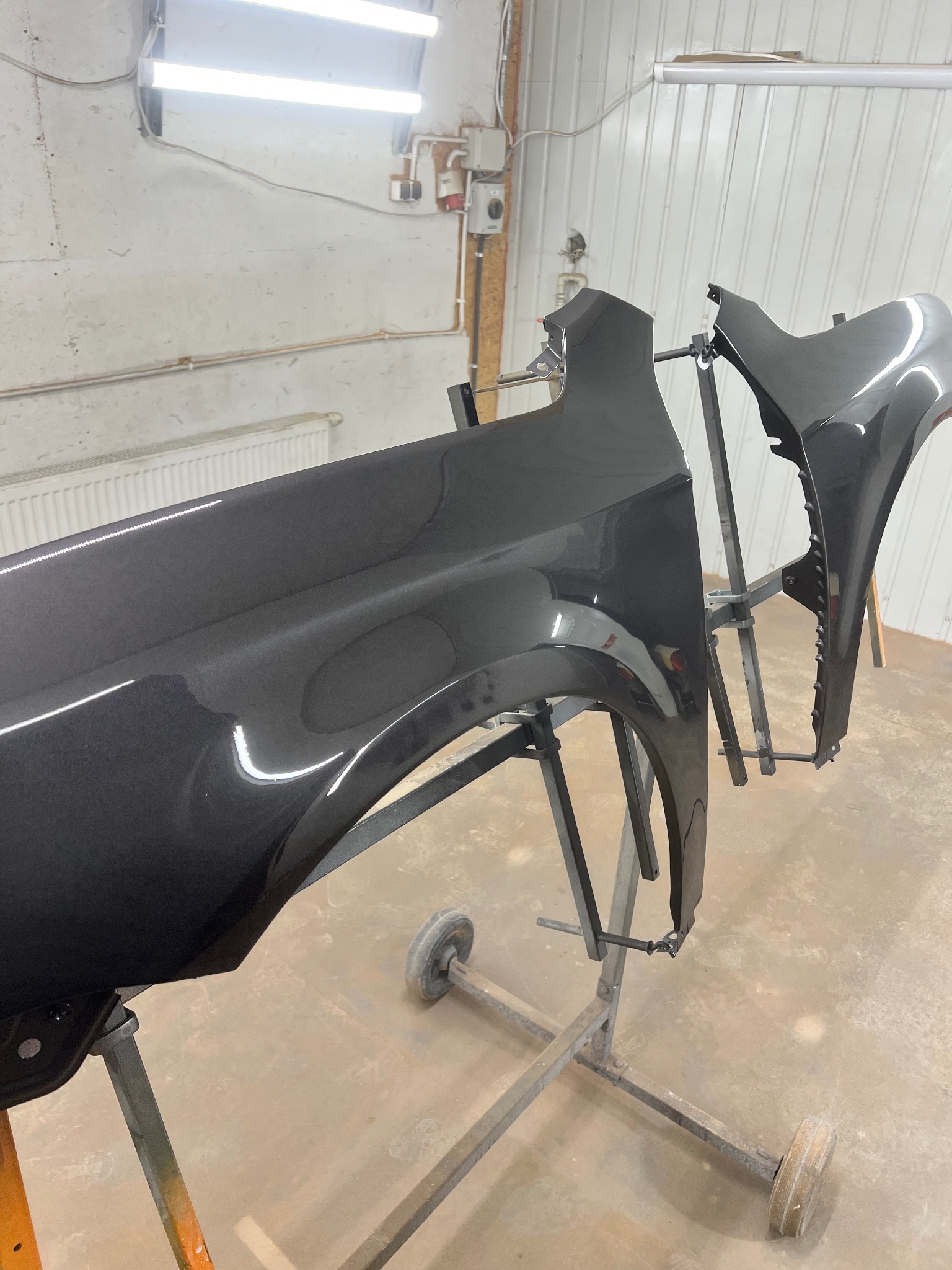 Błyszczące, pomalowane na grafitowo elementy karoserii samochodowej, zawieszone na metalowym stojaku w warsztacie lakierniczym, oświetlone lampami jarzeniowymi.