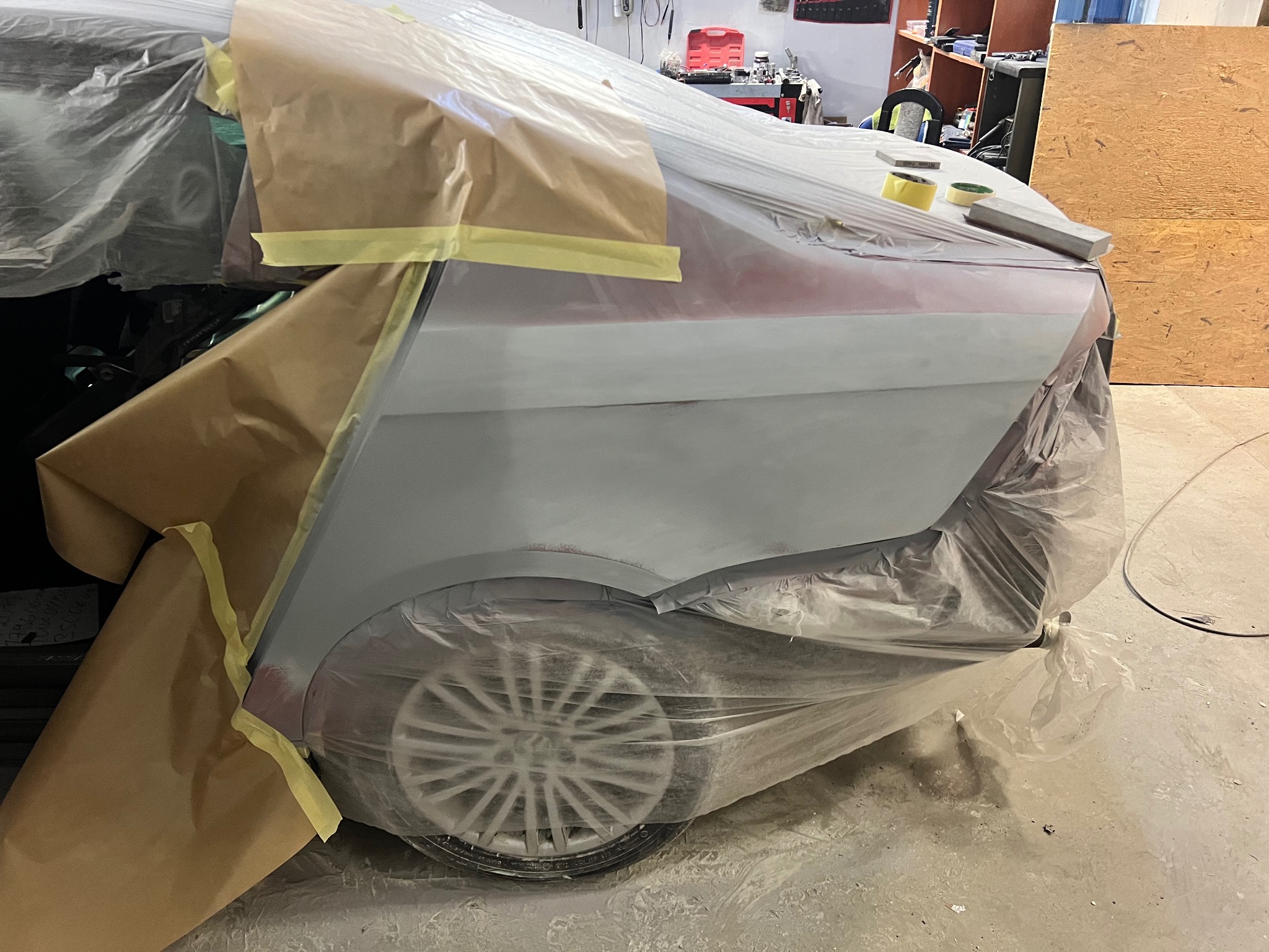 Przygotowanie do lakierowania auta w Krynicznie: zabezpieczony folią i papierem element karoserii po nałożeniu podkładu, widoczne koło. Etap prac blacharsko-lakierniczych.