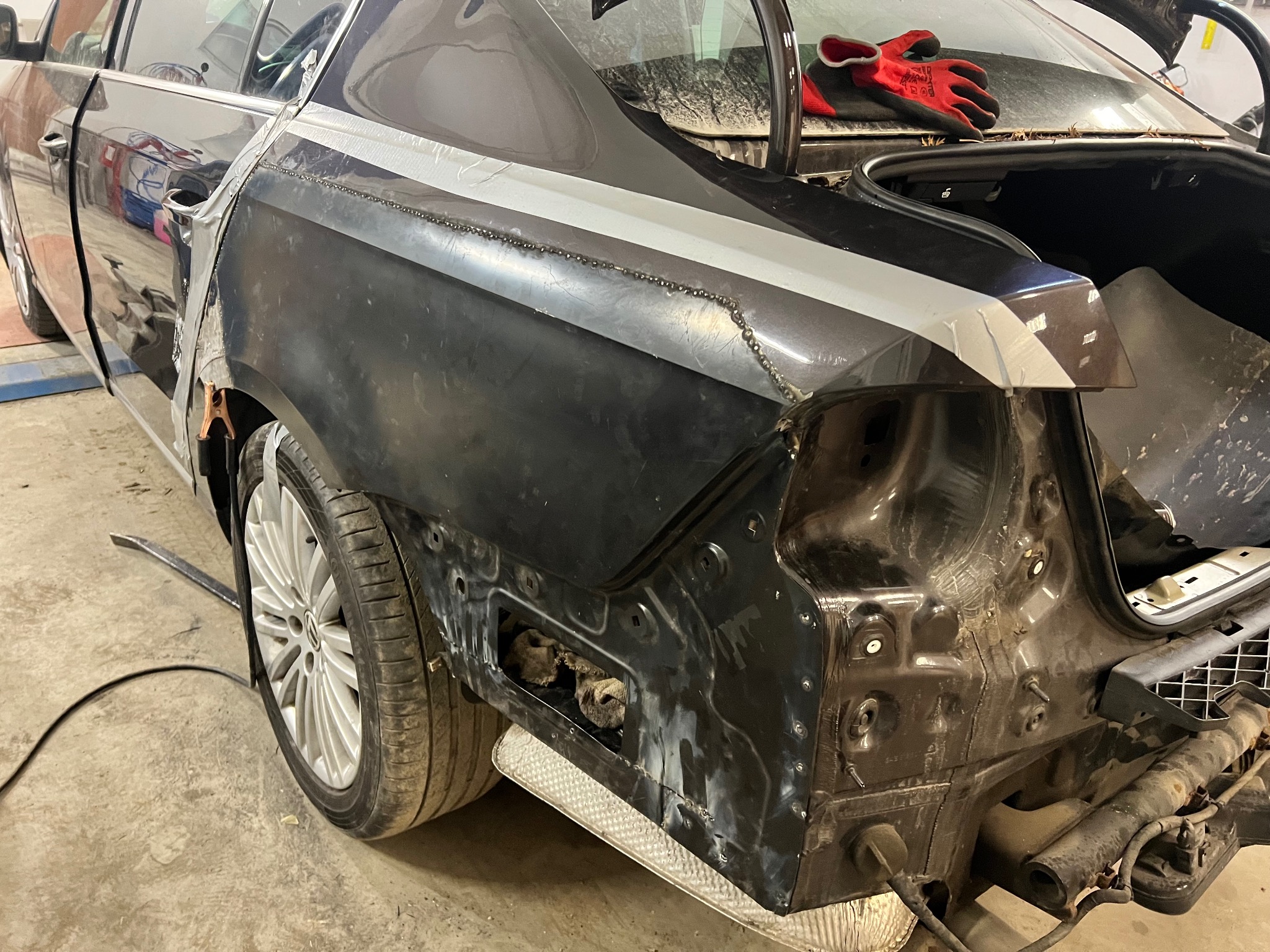 Częściowo zdemontowany, czarny samochód w warsztacie. Widoczne ślady spawania na karoserii, przygotowanie do lakierowania. Czerwone rękawice leżą na bagażniku.