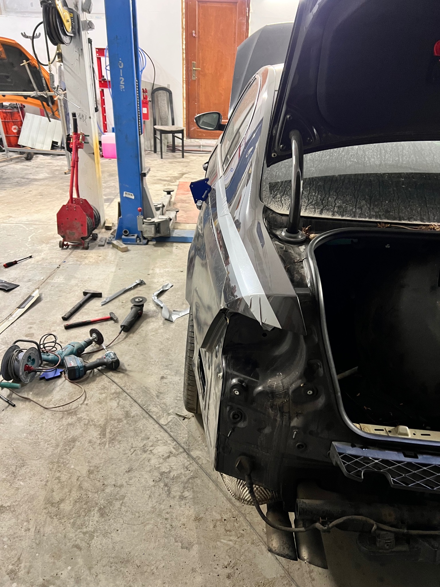 Uszkodzony tył auta w warsztacie, widoczne narzędzia i podnośnik. Błotnik w trakcie naprawy, oklejony taśmą. Otwarta klapa bagażnika.