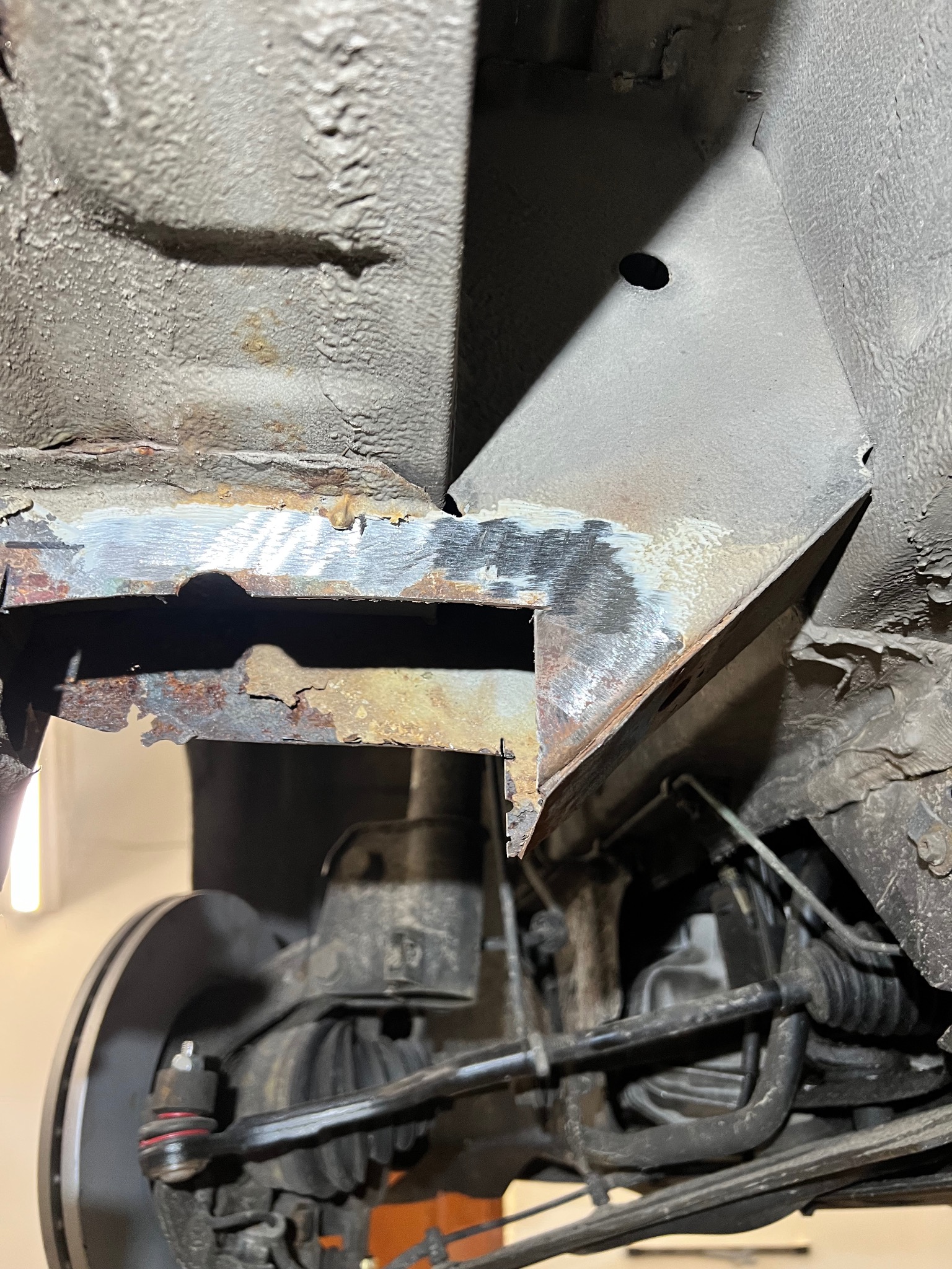 Fragment podwozia auta w trakcie naprawy blacharskiej. Widoczne elementy skorodowane i obszary po oczyszczeniu z rdzy, przygotowane do spawania. Ujęcie z dołu, oświetlenie warsztatowe.