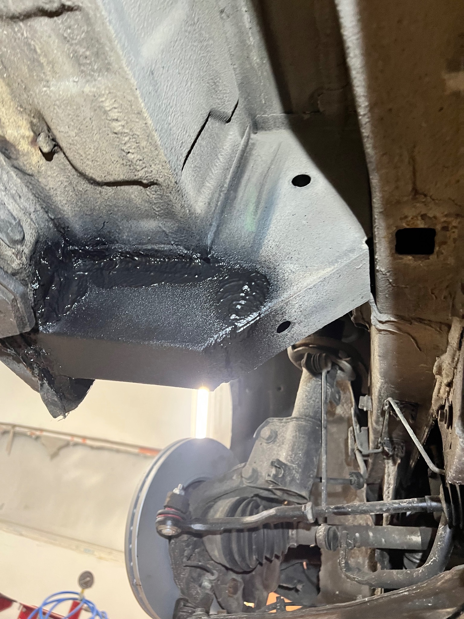 Podwozie samochodu po konserwacji antykorozyjnej, widoczne świeżo nałożone, czarne zabezpieczenie na elementach zawieszenia i podwozia. Ujęcie z dołu, w warsztacie.