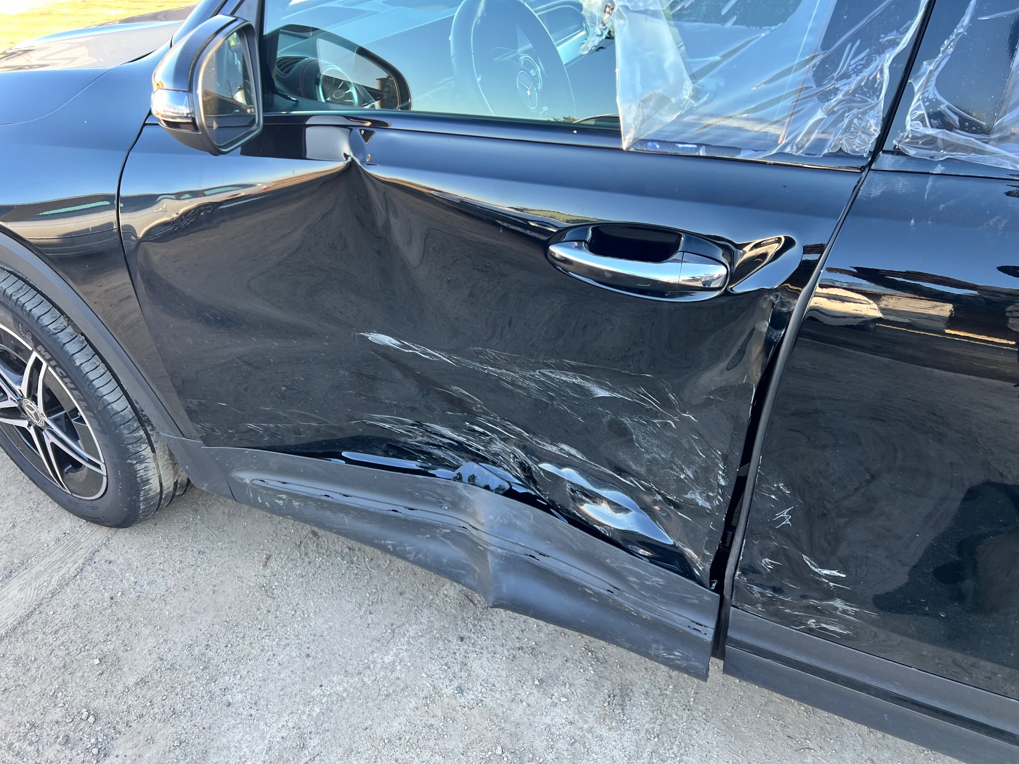 Uszkodzony bok czarnego samochodu z widocznymi wgnieceniami i zarysowaniami na drzwiach i błotniku, sugerujący potrzebę naprawy blacharsko-lakierniczej.