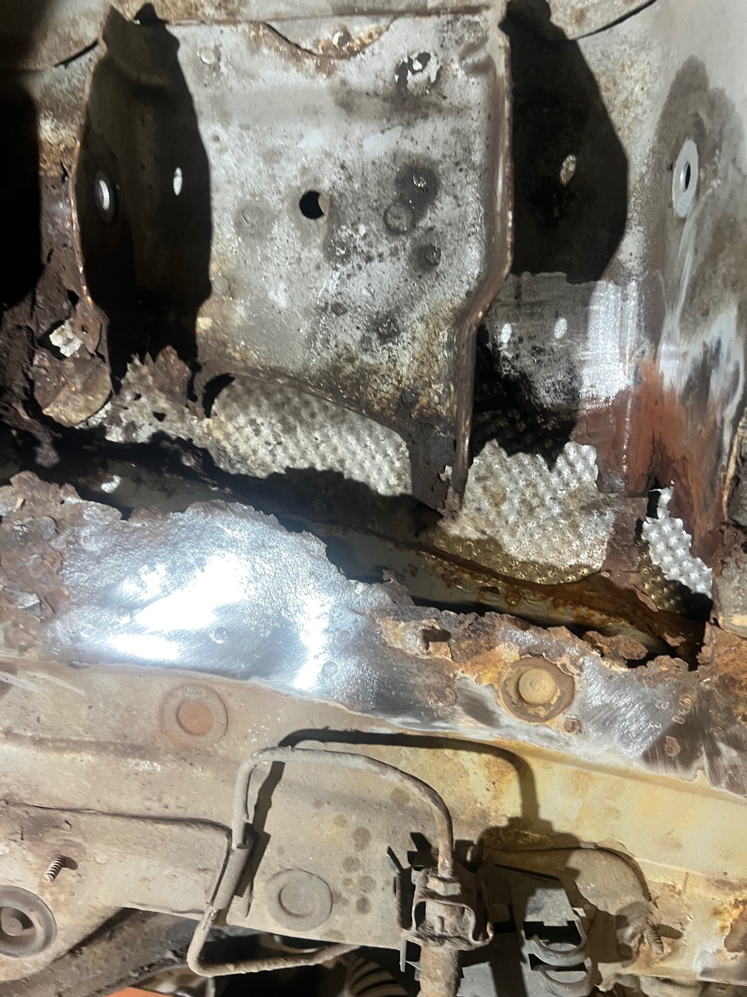 Zbliżenie na skorodowaną podłogę samochodu w trakcie naprawy. Widoczne ogniska rdzy i fragmenty oczyszczone do gołej blachy. Elementy zawieszenia w dolnej części kadru.