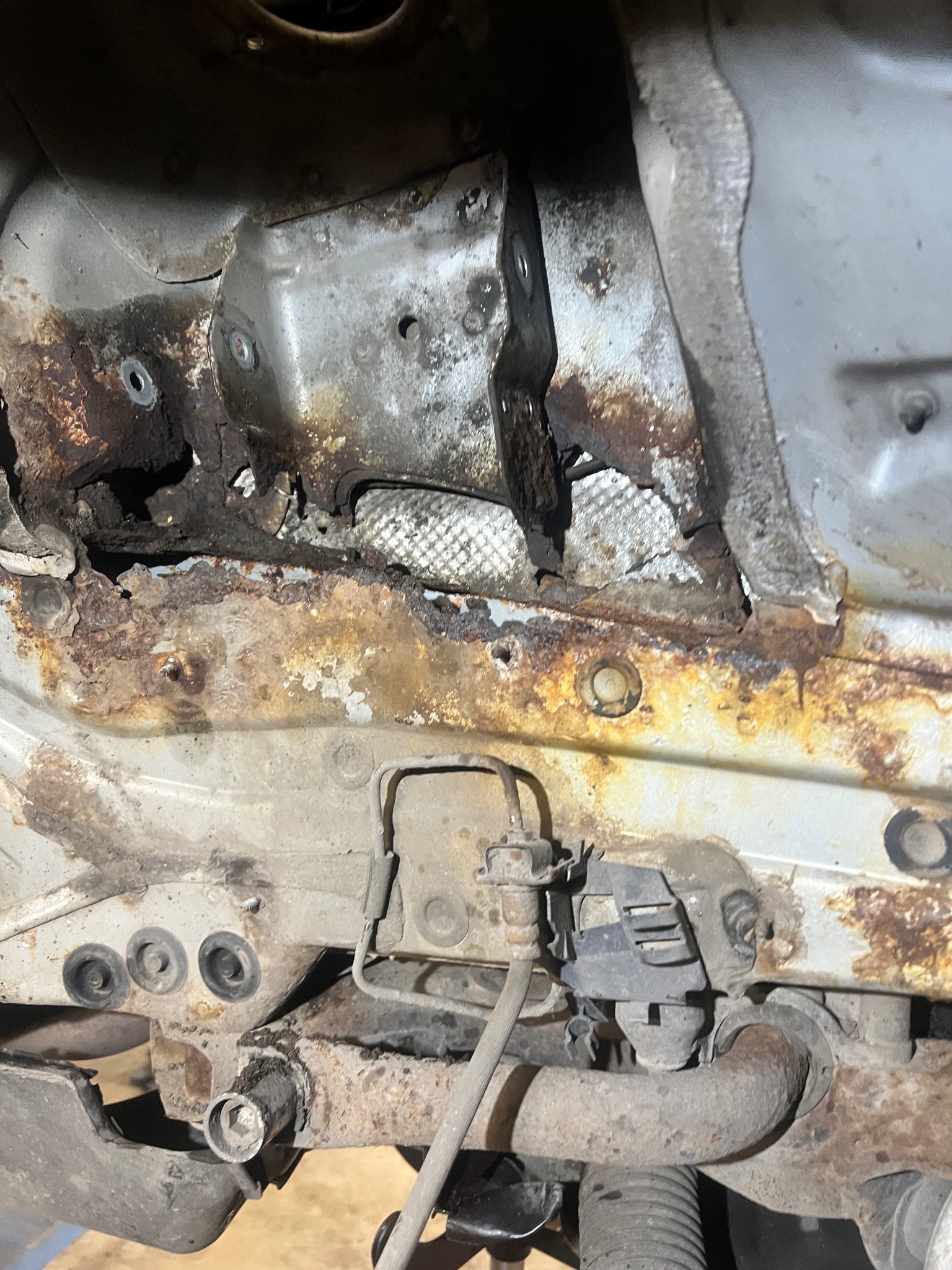 Zardzewiałe podwozie samochodu z widocznymi elementami zawieszenia i przewodami. Silna korozja z ubytkami materiału. Detal z bliska, ukazujący stopień zniszczenia.