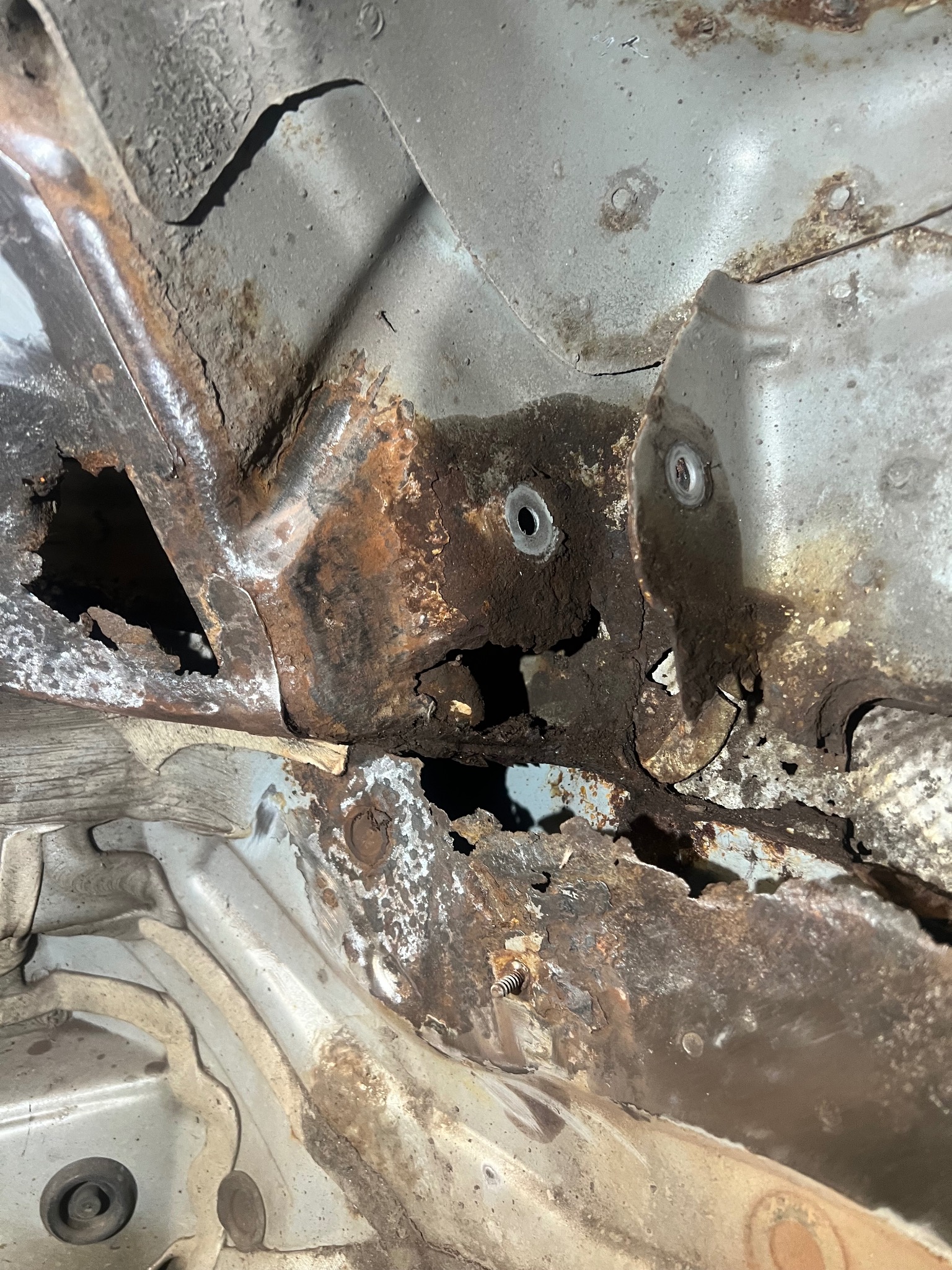 Zniszczona korozją podłoga samochodu z widocznymi dziurami i rdzawym nalotem. Fragment karoserii z otworami montażowymi i śladami napraw blacharskich.